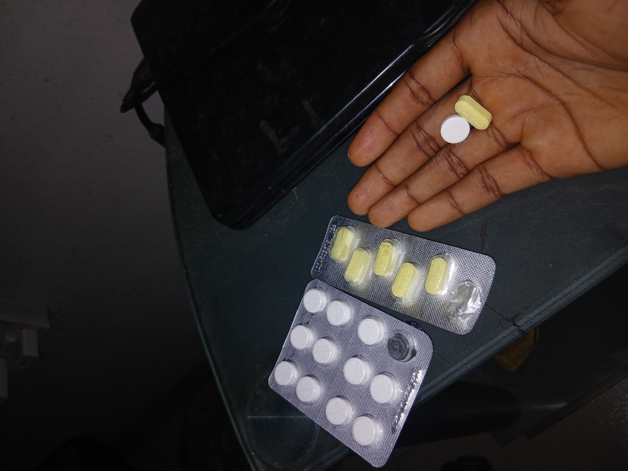 A close-up photograph shows a dark-skinned human hand holding a white round pill and a yellow oblong pill. Below the hand, two blister packs of pills rest on a dark green surface. One blister pack contains white round pills, one of which is missing. The other blister pack contains yellow oblong pills, one of which is also missing. A black object is partially visible in the upper left corner of the frame. The location is unknown.