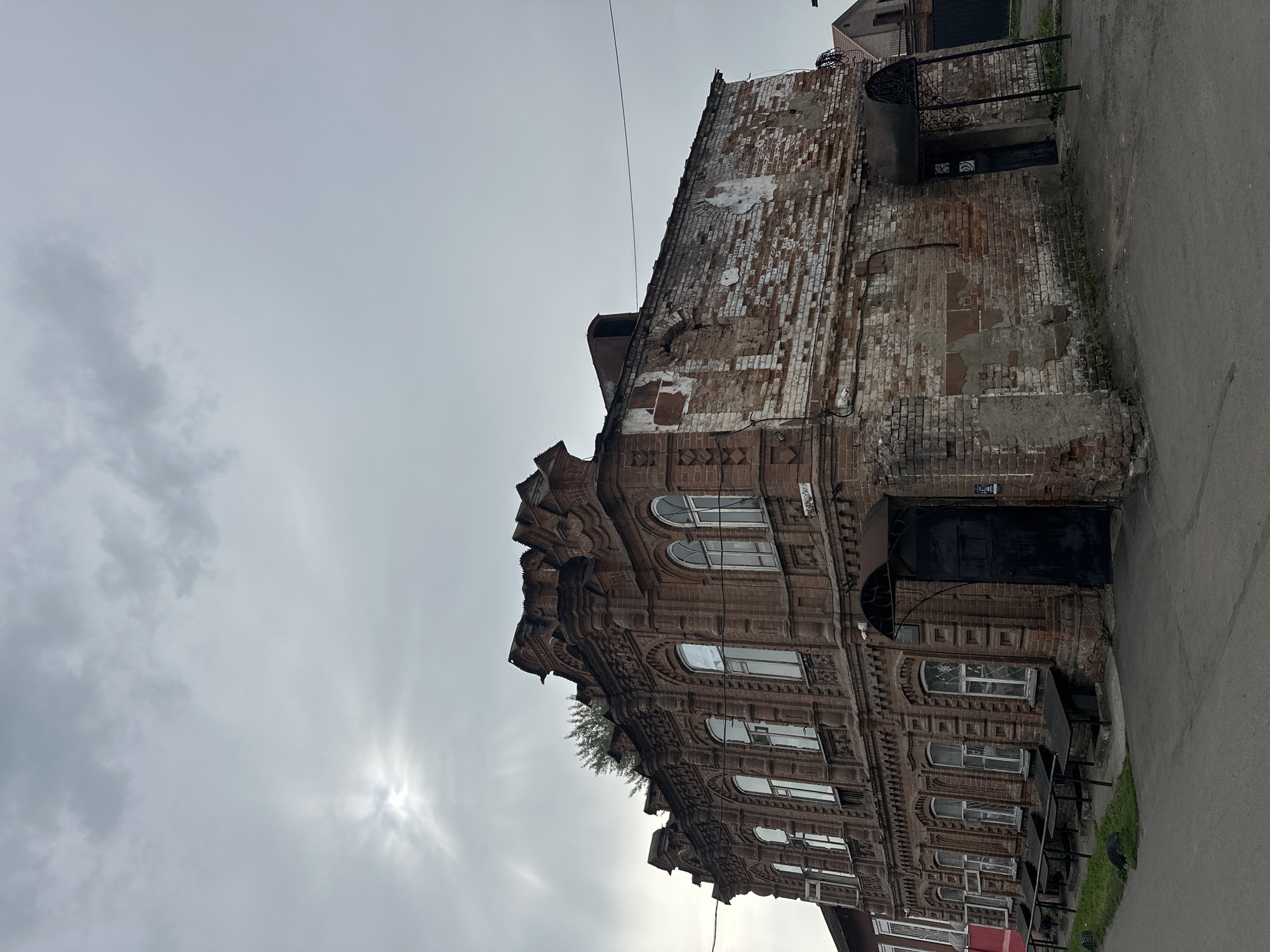 An exterior, medium shot shows a corner view of a two-story, weathered brick building in Biysk, Russia, under an overcast sky with the sun peeking through the clouds. The building's facade is intricate, with decorative brickwork along the roofline and around the arched windows on the upper floor. The lower portion of the building, particularly on the right side, shows significant deterioration, with exposed brick and peeling plaster. Two dark awnings cover doorways. In front of the building on the left, a paved walkway runs alongside a grassy area, with several dark benches or tables. The sky is filled with thick, grey clouds, with the sun creating a diffused glow.