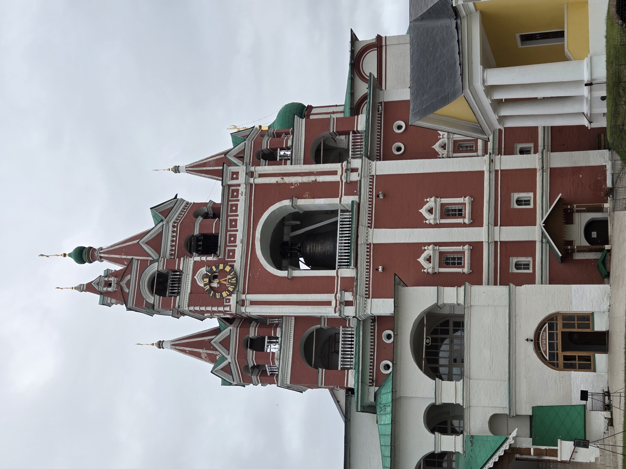 The image depicts the Bell Tower of the Savvino-Storozhevsky Monastery in Zvenigorod, Russia, under a cloudy sky. The tower is a prominent red brick structure with white accents, featuring multiple levels with arched openings, balconies, and ornate detailing. A large bell is visible within one of the openings. The clock face on the tower is golden with Roman numerals. Green domes and roofs are integrated into the architecture. In the foreground, a portion of a white building with arched windows and a wooden entrance is visible, labeled with the Cyrillic text "СВЯТАЯ ВОДА". To the right, a smaller yellow building with white columns and a dark roof is partially visible. The ground is paved with light-colored tiles and bordered by a dark metal fence and green grass. No people are visible in the image.