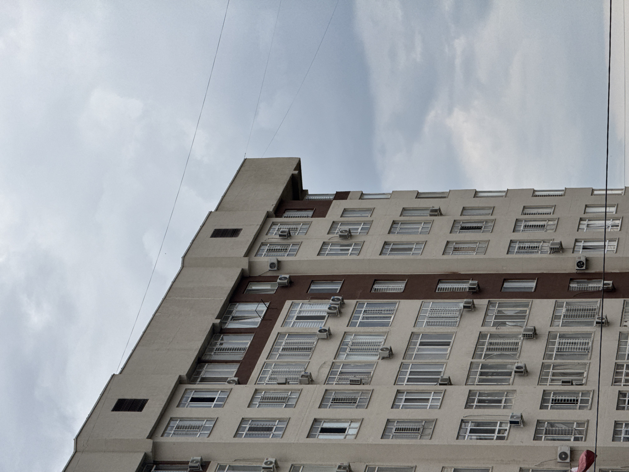 An exterior, medium shot captures the side of a multi-story apartment building against a cloudy sky in an urban environment. The building features numerous windows with white frames, some with bars, and many have air conditioning units mounted below them. The facade is predominantly a light beige color, with a section of dark brown accentuating some of the windows. A red object, possibly a banner or balloon, is partially visible on the lower left side of the building. Power lines are visible stretching across the upper portion of the image.