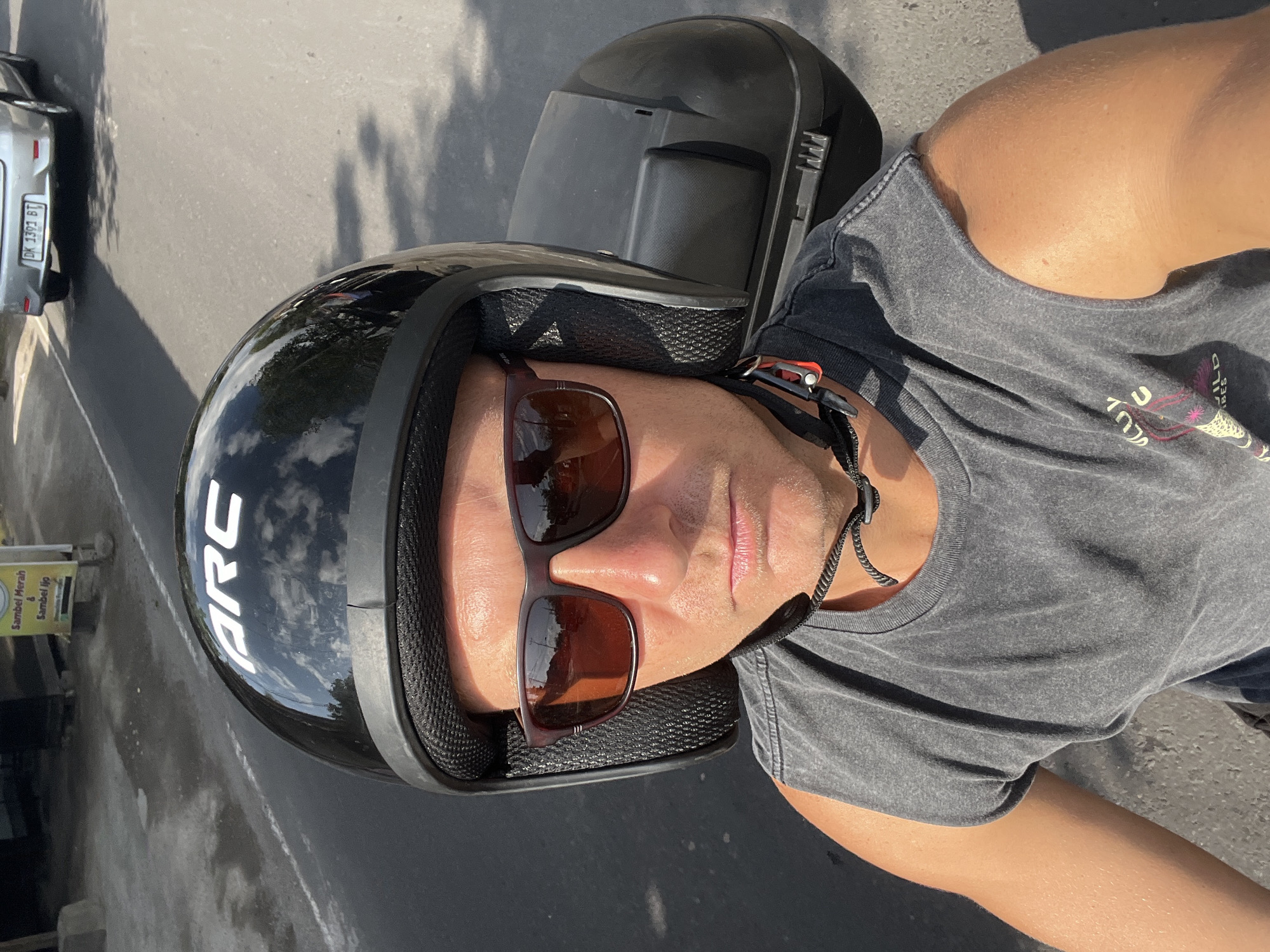 This media file shows a close-up, overhead view of a person wearing a black motorcycle helmet and sunglasses. The person is also wearing a grey t-shirt. The helmet has a white "ARC" logo on the side. The scene appears to be outdoors, on a road, with a sliver of a silver car visible on the left side of the frame. There is also a yellow sign with text in a foreign language visible in the background. The lighting suggests it is daytime, possibly late afternoon, given the shadows. The overall tone is casual and likely depicts the person preparing for or in the midst of a motorcycle ride in Sukawati, Indonesia. The visible text on the sign reads "Sambel Merah & Sambel Ijo". The license plate on the car shows "DK 1393 BT".