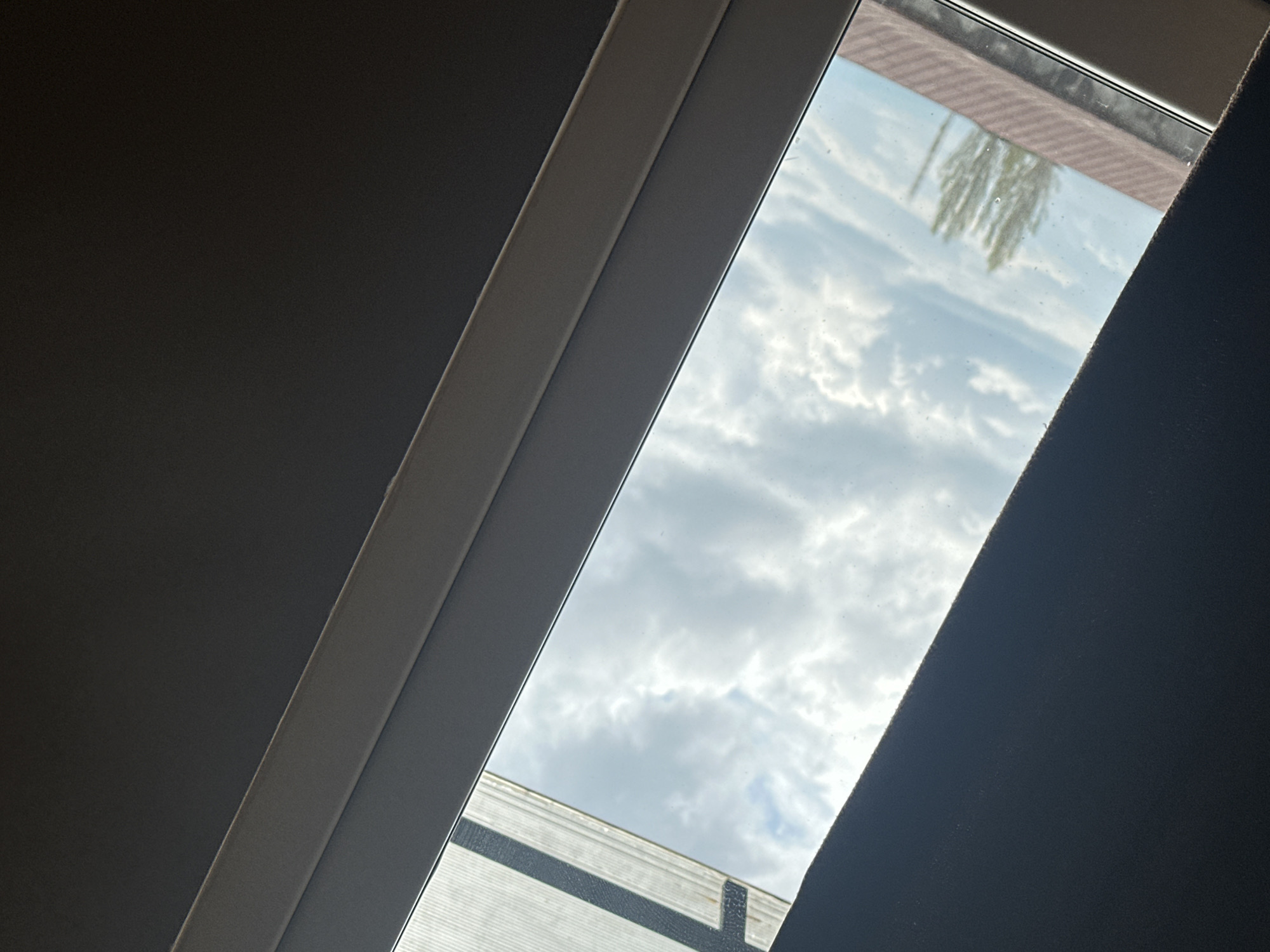 A low-angle shot looking up through a window reveals a partially cloudy sky and the top of a tree. Part of a black curtain is seen on the right side of the image. The photograph appears to be taken indoors, with sections of the ceiling visible near the top and bottom of the window frame.