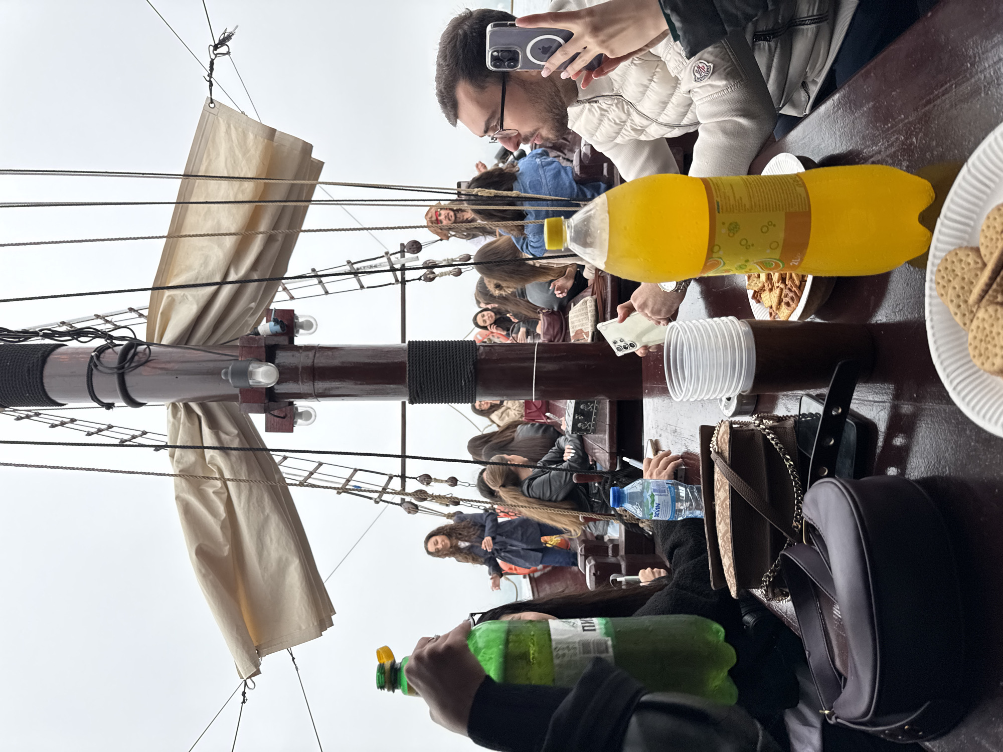 Captured on a boat, possibly on the Black Sea, is a daytime scene featuring numerous people, including one man actively taking a photo with his smartphone. The photo is taken from a low vantage point, looking upward at the mast and sails of the boat. Passengers are seated along a long table. Beverages, including bottles of orange juice and water, and snacks such as cookies and mixed nuts, are present on the table. The sky is overcast. The specific location in Varna, Bulgaria, is indeterminable from the image.