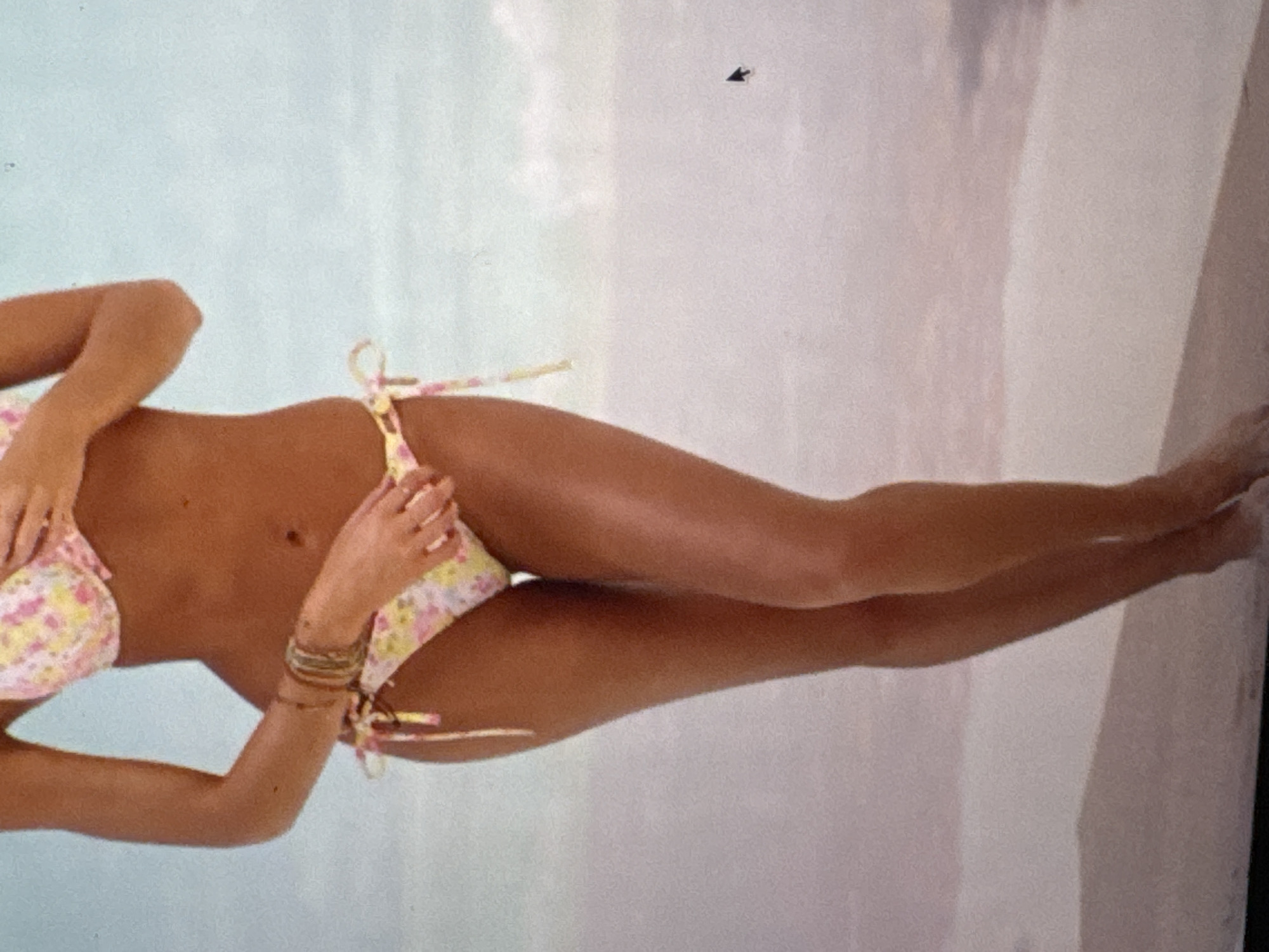 This media file shows a close-up, vertically oriented shot of a person's torso and legs. The subject is wearing a floral bikini with pink and yellow flowers on a white background. The bikini ties at the sides of the hips. The person has tanned skin and is adorned with several bracelets on their left wrist, which appear to be made of beads and thin metal bands. The background is a softly blurred pastel blue and pink, suggesting a beach or water setting, potentially at Kuta Selatan, Indonesia. The lighting is soft and diffused, implying daylight. There is a small, dark, arrow-shaped object visible in the upper right quadrant of the image, which may be an insect or a blemish on the original photograph. The overall impression is one of leisure and relaxation in a tropical environment.