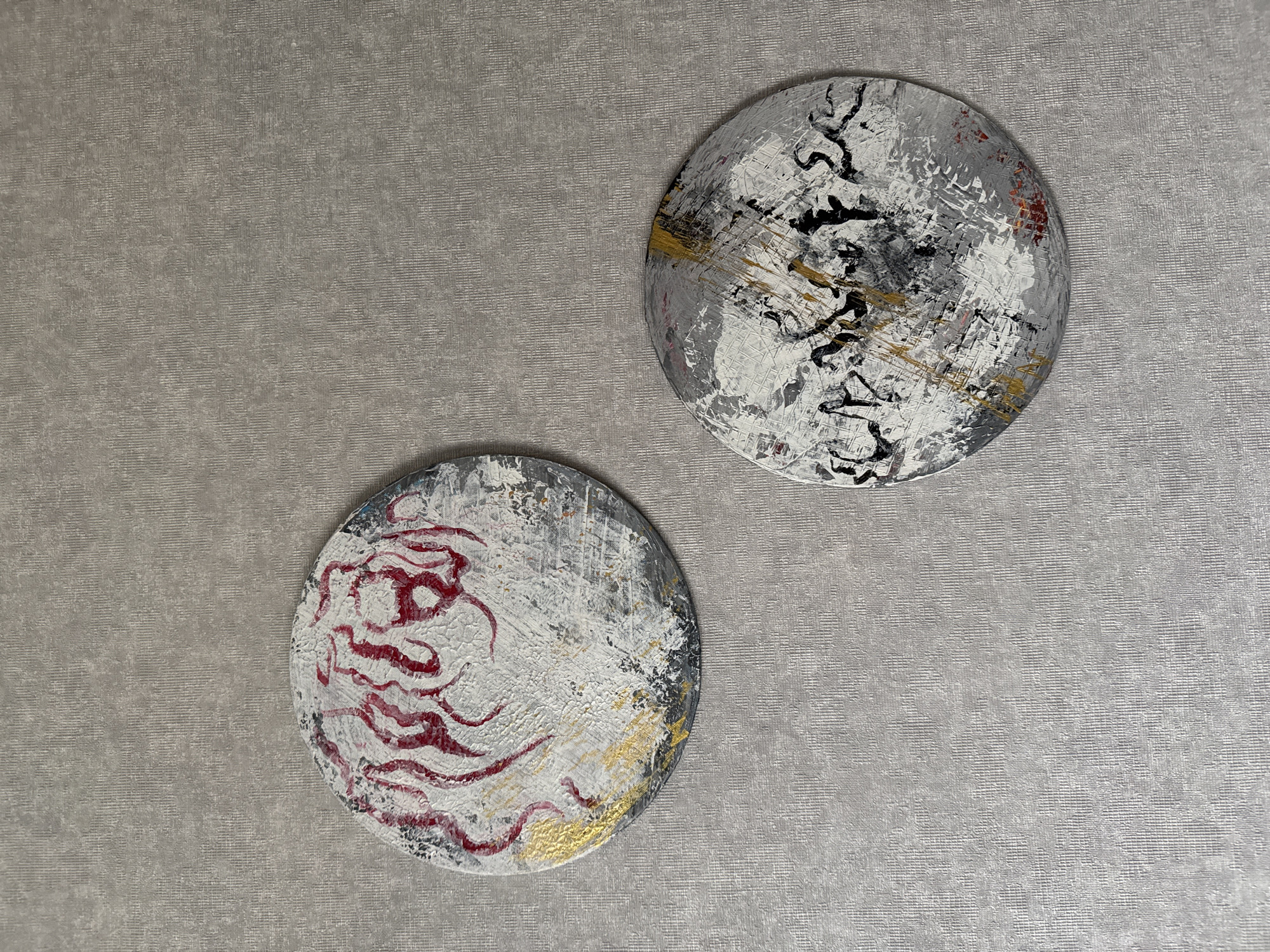 The image presents two circular abstract art pieces displayed on a textured wall. The wall exhibits a uniform light grey-beige color with a subtle, fine horizontal ribbed or woven pattern.
The two artworks are positioned diagonally across the frame; the upper right circle is slightly larger and situated higher than the lower left one. Both pieces share a consistent abstract style, characterized by a highly textured, layered application of paint primarily in white, various shades of grey, and black.
The upper right artwork features a dominant monochrome palette. It showcases thick, impasto-like white paint with noticeable brushstrokes, revealing underlying grey tones. Prominent, stylized, calligraphic-like black strokes or abstract shapes resembling intertwined forms are spread across its surface. Accents of metallic gold and faint hints of red or dark pink are also visible, particularly towards the periphery.
The lower left artwork similarly utilizes a base of textured white, grey, and black paint. Its central motif consists of flowing, organic, and highly textured red (or deep burgundy) abstract shapes, which could be interpreted as tendrils, flames, or abstract script. Metallic gold paint accents are present towards the bottom edge, complementing the cooler tones.
No activity or event is depicted; the image is a static representation of interior decor. The lighting suggests an indoor setting. There is no clearly legible text on either artwork, only abstract marks that evoke stylized characters or forms. The scene conveys a modern and artistic ambiance, characteristic of contemporary interior design.