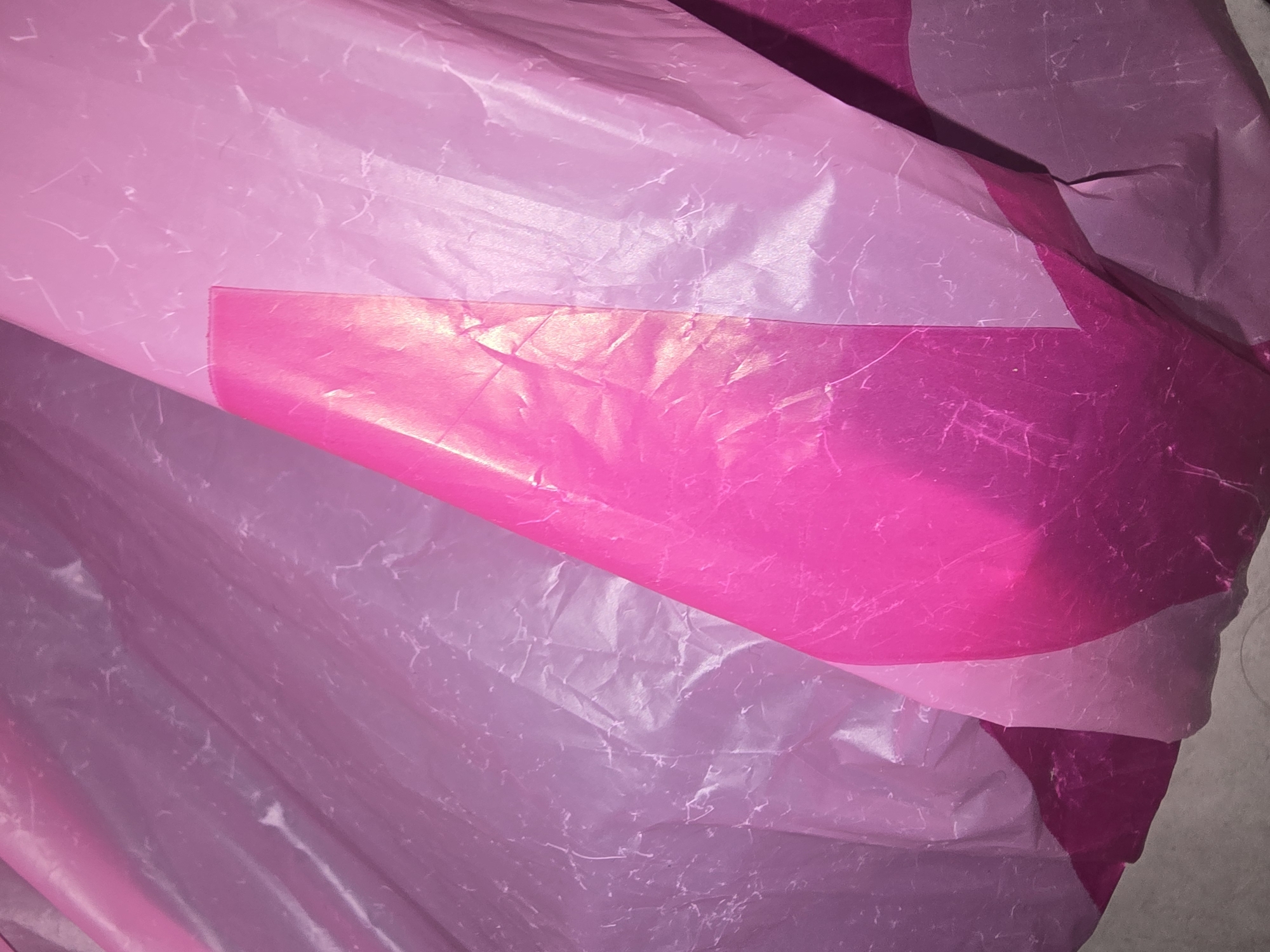 The image shows a close-up, abstract view of layered pink and purple translucent plastic sheeting. The material is crinkled, with visible creases and some faint linear markings that suggest a fibrous texture or a pattern within the plastic. The lighting creates highlights and shadows, emphasizing the dimensionality of the draped material. There are no discernible features to indicate a specific location in Ulan-Ude, Russia, nor are there any people or specific objects visible. The scene is purely a textural and color study of the plastic.