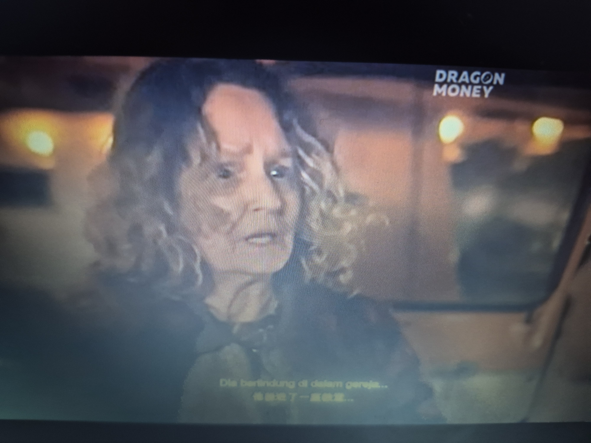 The image is a still from a screen, possibly a television or monitor, displaying a scene with an older person with curly hair, looking towards the left of the frame. The lighting is dim, with warm tones coming from the background, suggesting artificial lights. In the upper right corner of the screen, the text "DRAGON MONEY" is visible in white font. Below the person, there are subtitles in two lines, the first line appearing to be in Malay or Indonesian ("Dia bertindung di dalam gereja...") and the second line in Chinese characters. The scene appears to be indoors, with an out-of-focus background that includes soft, glowing lights. There is no discernible information in the image that would indicate a specific location within the city of Sandwell, United Kingdom.