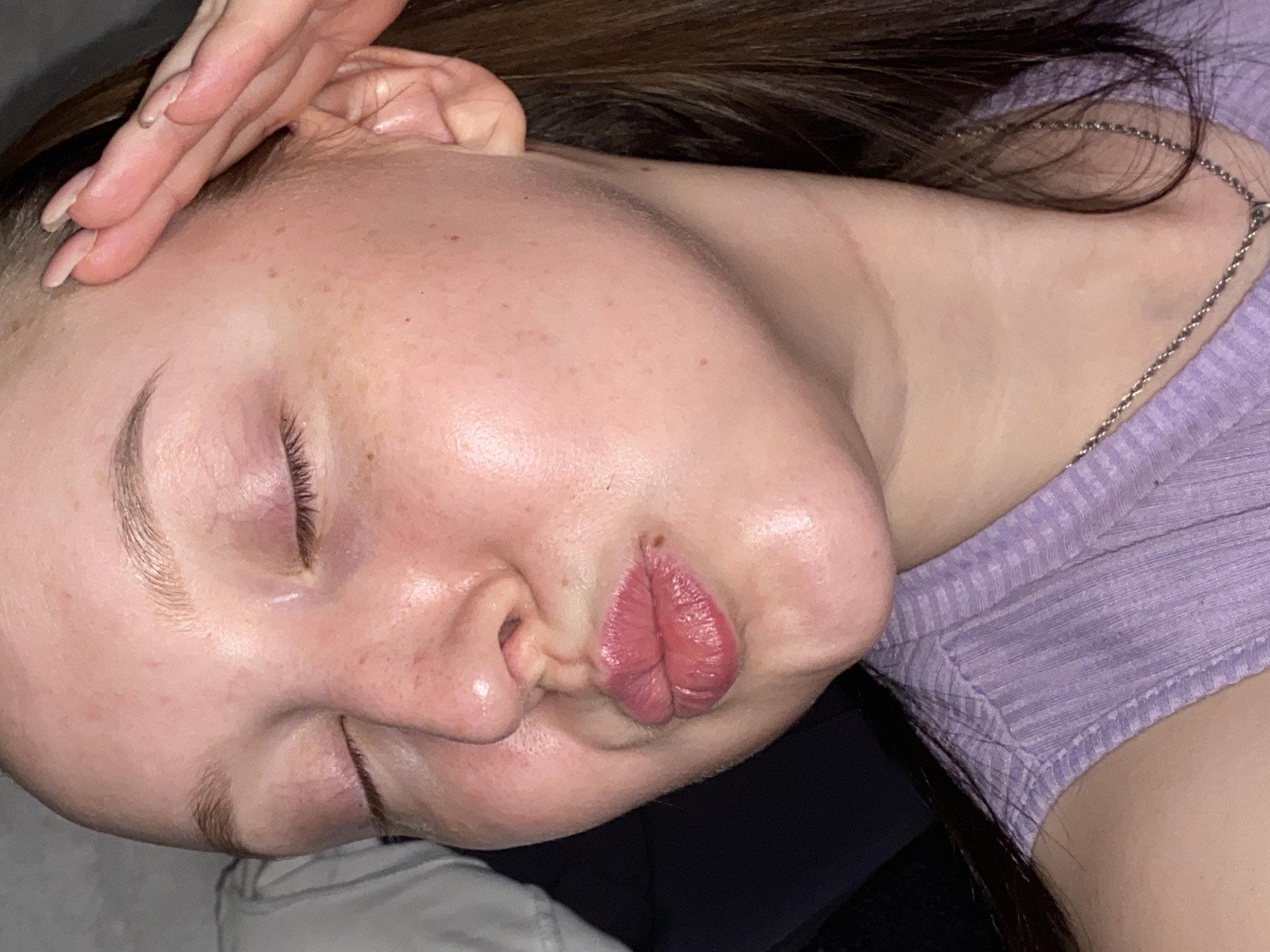 A close-up, angled shot shows a person, likely female, lying down with her eyes closed and lips pursed in a kissing gesture. Her face is pale with some freckles visible across her forehead and cheeks, and her eyebrows are well-groomed. There is a slight sheen on her skin, possibly from moisturizer or natural oils. One hand with light-colored fingernails rests gently on her temple, near her dark brown hair which is spread out behind her head.
She is wearing a ribbed, light purple top and a thin silver chain necklace is visible around her neck. Her expression, combining closed eyes and pursed lips, suggests a moment of playful relaxation or a posed gesture. The lighting appears soft and artificial, indicative of an indoor setting. The scene is set in Chaikovsky, Russia. There is no visible text in the image.