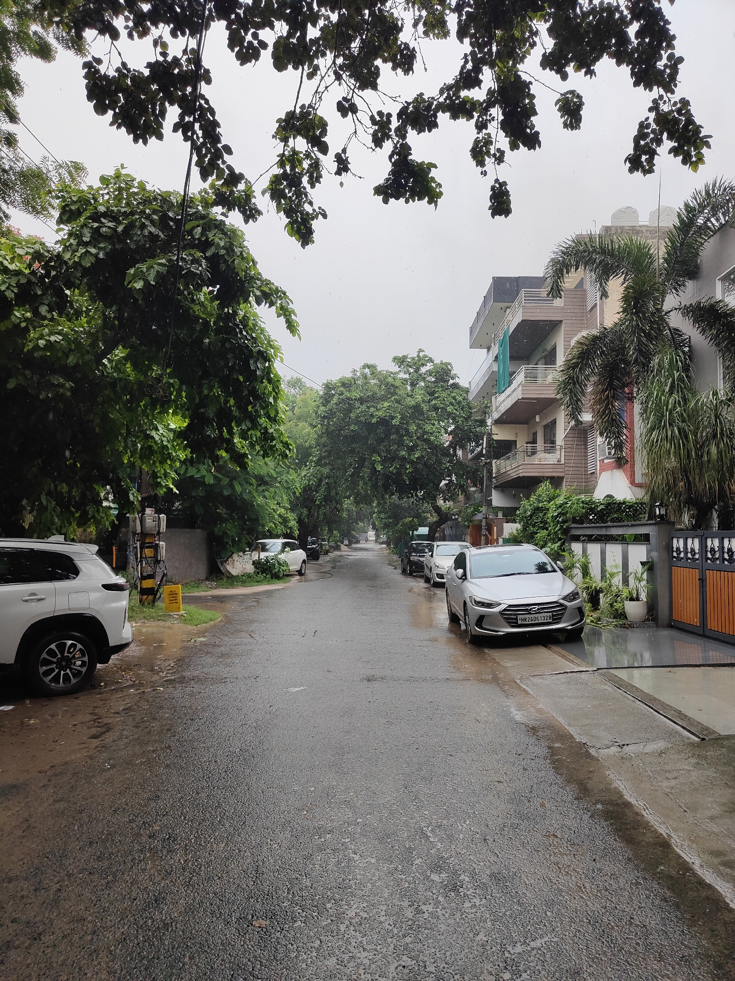 The image depicts a street scene in Gurgaon, India, during what appears to be rainfall. A wet asphalt road stretches into the distance, lined by residential buildings and trees. On the left side of the road, a white SUV is parked near a utility pole with visible electrical meters. Further down the street, more vehicles are parked, including a silver sedan with a license plate that reads "HR26DG1328." Green trees and foliage are abundant on both sides of the street, with some branches overhanging the road from the top of the frame. The sky is overcast, and the overall atmosphere suggests a dreary or rainy day. The buildings are modern multi-story structures with balconies and balconies, typical of residential areas in the city. A wooden and metal gate is visible on the right side of the road, leading to a property. The ground is wet and reflective, indicating recent or ongoing precipitation.