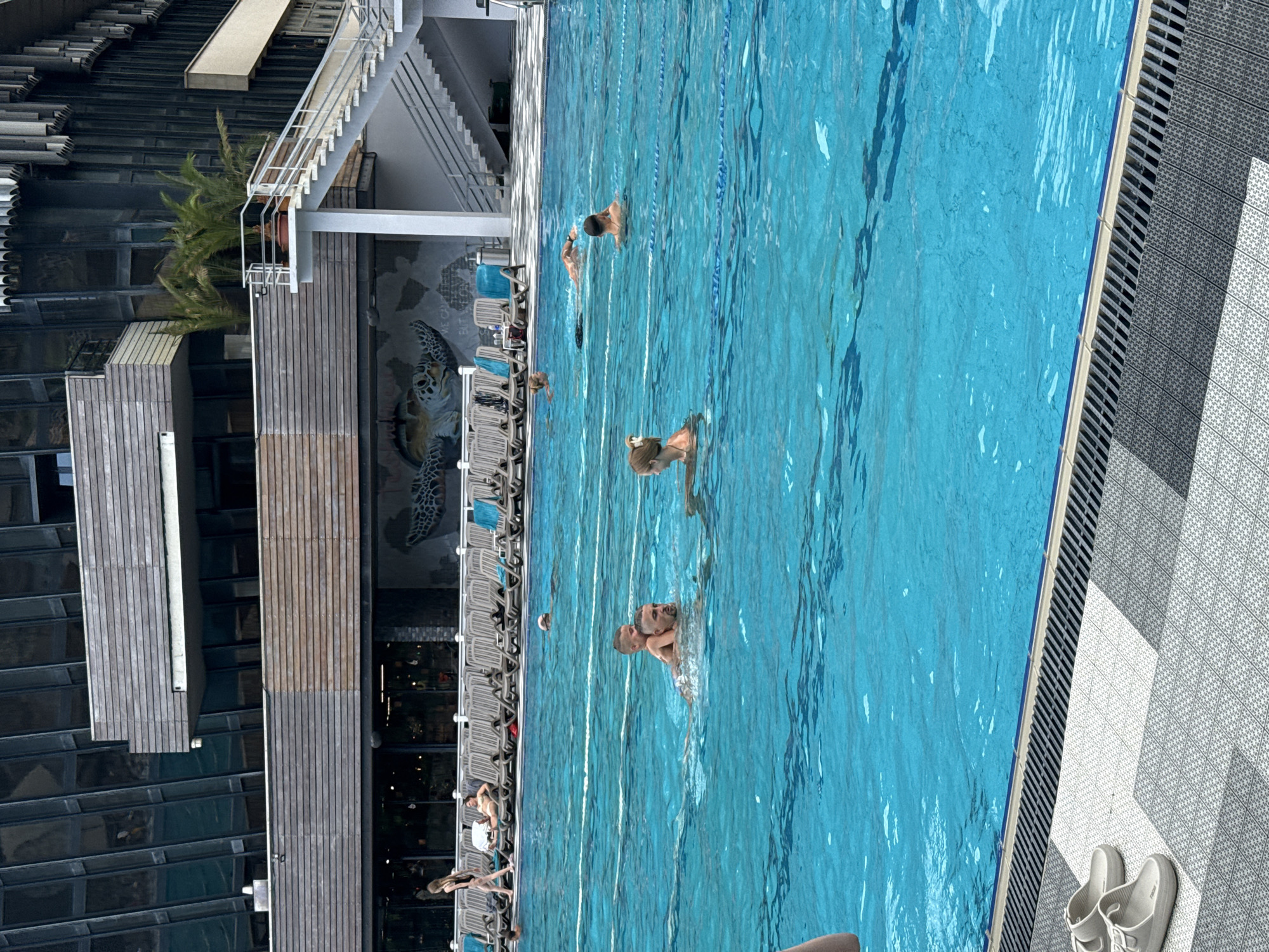 The image captures a bright, sunny day at an outdoor swimming pool, likely part of a hotel or resort in Yalta, Ukraine. In the foreground, a man with a child on his back is swimming in the clear blue water. To the right, a woman with blonde hair pulled back is also swimming. In the middle distance, another person is swimming. Further back, a woman with blonde hair is walking along the poolside, and a man is lounging on a chair. The background features a large building with a modern architectural design, characterized by glass windows and wooden accents. A mural of a turtle is visible on the building's exterior. The poolside area is furnished with numerous lounge chairs and a staircase leading up to a higher level of the building. The water appears inviting, with lane markers visible beneath the surface. A pair of light-colored flip-flops rests on the tiled edge of the pool in the lower left corner of the image. The overall atmosphere is one of relaxation and recreation.