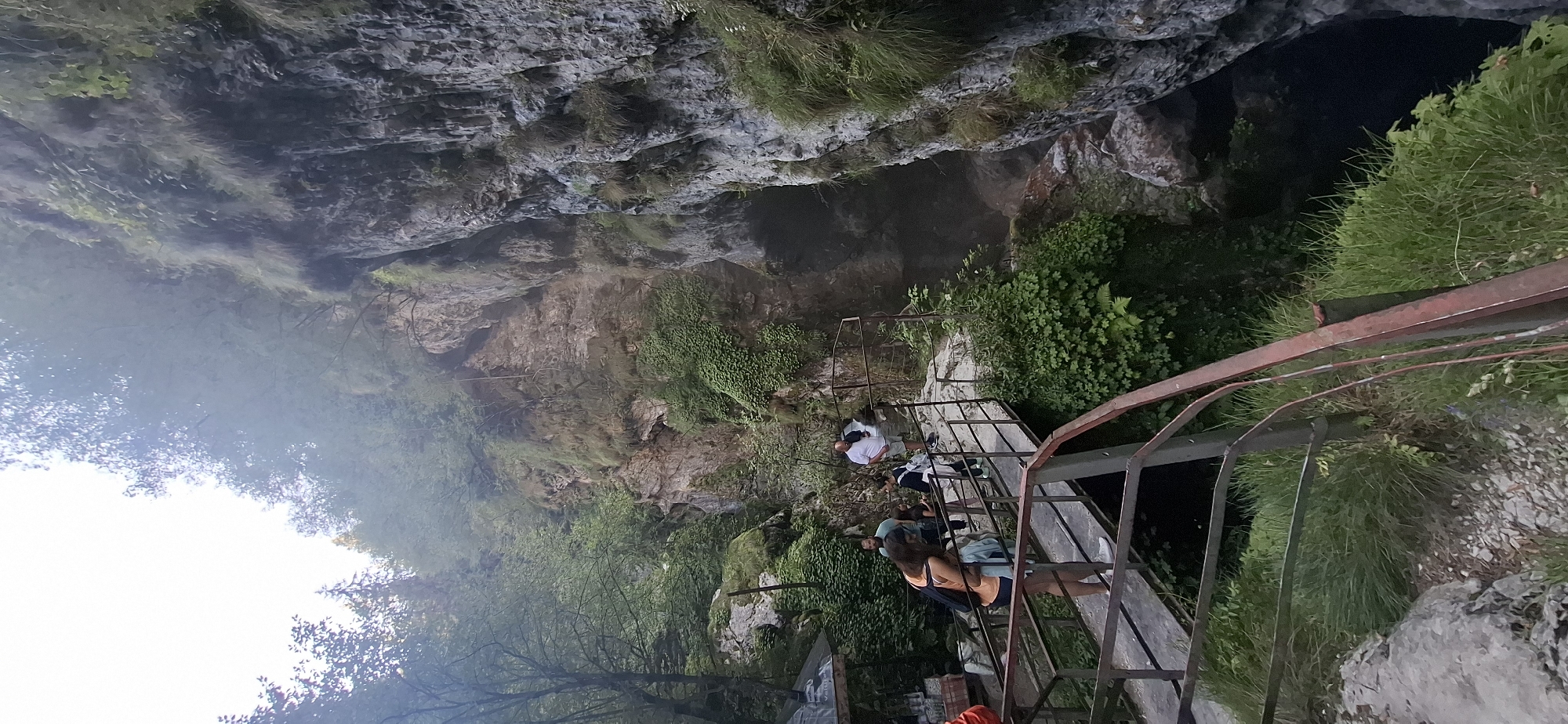 This is a vertical, eye-level, full shot capturing a view of the Trigrad Gorge in Trigrad, Bulgaria. The scene features a narrow metal walkway, painted red and dark gray, with people on it, overlooking a deep, dark chasm to the right. The walkway is lined with a rusty red metal railing.
On the walkway, there are at least five individuals. In the foreground on the left, a woman with long brown hair is wearing an orange tank top and dark shorts, carrying a light blue item over her shoulder. Behind her, a person in a light blue shirt and dark pants stands. To their right, two people are facing away from the camera. One person, wearing a white polo shirt and khaki shorts, is standing near a railing, looking into the gorge. Another person in a dark shirt and backpack stands beside them.
The gorge walls are steep and rocky, covered in lush green vegetation. The sky above is overcast and bright, with some trees visible on the left side of the image. The overall impression is of a natural tourist attraction with a constructed pathway for visitors to observe the dramatic landscape.