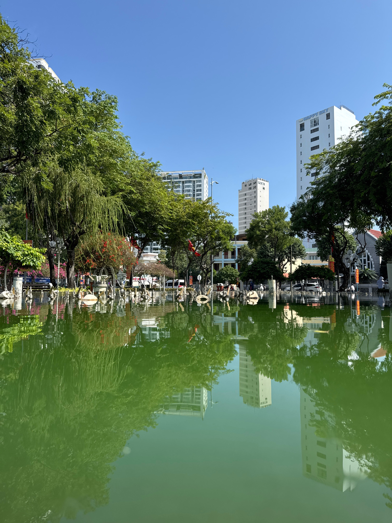 This is an outdoor, low-angle, eye-level shot of a green pond in Nha Trang, Vietnam. The pond reflects the surrounding environment of trees and buildings, creating an almost surreal effect. In the foreground, the vibrant green water dominates the frame, showing distorted reflections of the trees and architecture. Scattered across the pond are various decorative elements, some appearing to be stone or metal sculptures, resembling swans and other artistic features.
In the midground, a park-like setting is visible. Lush green trees, including a weeping willow on the left, provide shade and frame the scene. Interspersed among the trees are what appear to be lampposts, decorative archways, and various structures. Some red and yellow flags are also visible, suggesting a possible celebration or decorative theme. A few people are scattered throughout the park, some seated, some standing, appearing to be enjoying the public space.
In the background, modern high-rise buildings rise against a clear blue sky. One building, prominently featured on the right, has the words "HAPPY SKY" visible on its facade. The buildings are a mix of white and light gray, reflecting in the pond below. The overall impression is one of a serene urban oasis, where nature and architecture coexist, with the pond serving as a central reflective element. The lighting suggests it is daytime, with clear skies and ample sunlight.