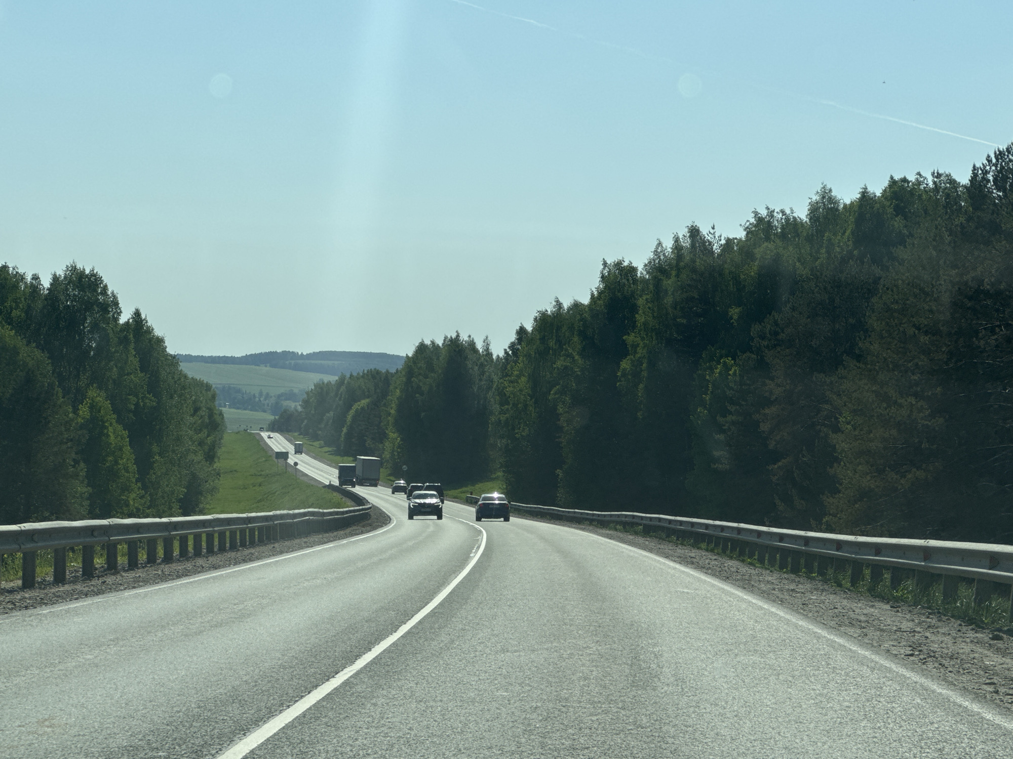 The image shows a two-lane asphalt road with a white dividing line, curving through a landscape of trees and hills. A metal guardrail runs along the left side of the road. Several vehicles, including cars and trucks, are visible in the distance. The sky is light blue and clear. The scene is located near Selishcha, Russia, suggested by the environmental clues.