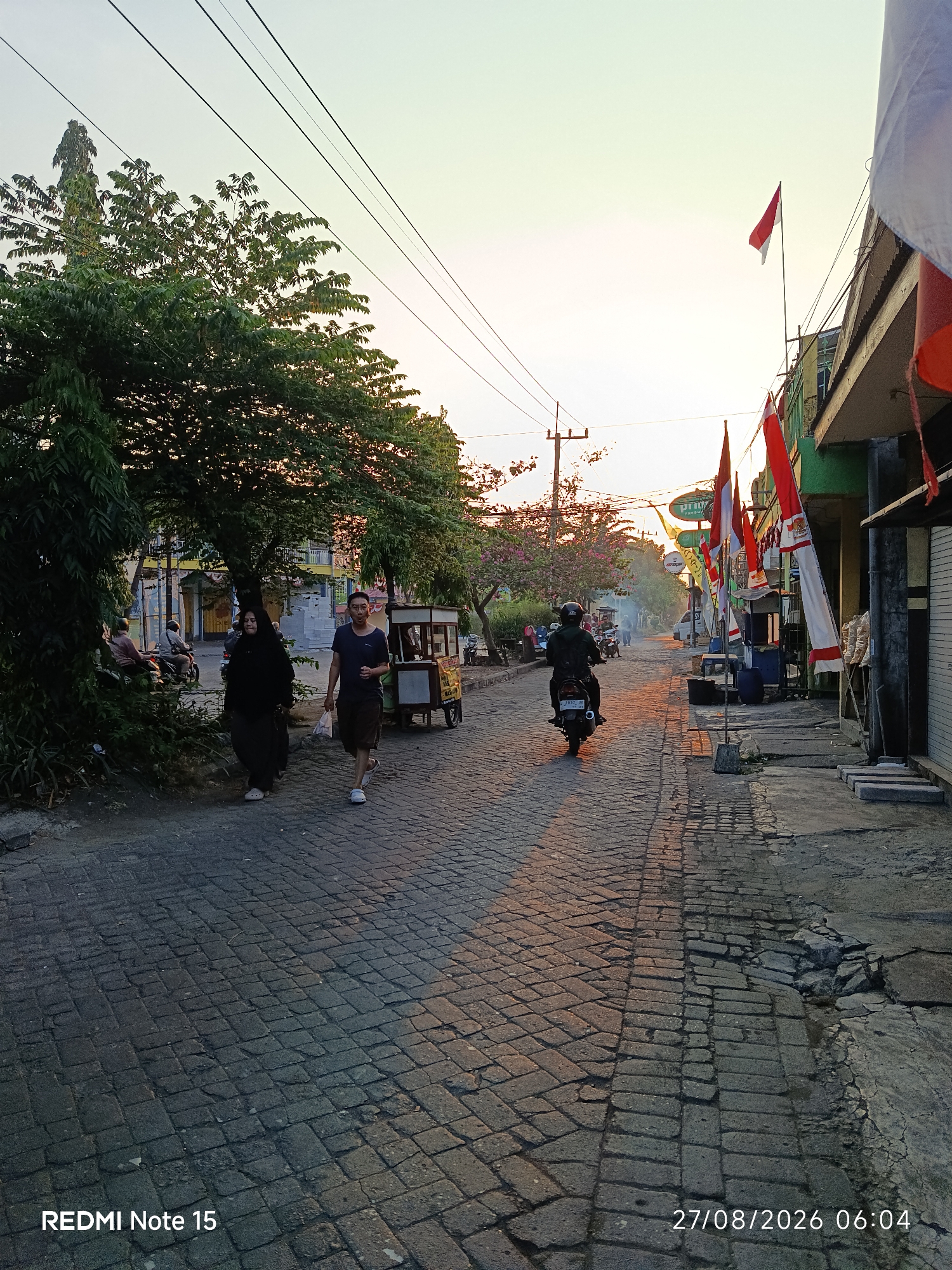 The image depicts a street scene in Keboansikep, Indonesia, during what appears to be early morning or late afternoon given the soft lighting and long shadows cast on the cobblestone road. Several Indonesian flags are prominently displayed along the right side of the street, indicating a possible local celebration or commemoration.
In the foreground, a man and a woman are walking on the cobblestone street. The woman is wearing a black hijab and abaya, while the man is in a dark t-shirt and shorts. To their left, a food cart is parked on the side of the road. Further down the street, a person is riding a motorcycle, heading away from the camera. The street is lined with buildings on the right and trees and more buildings on the left. The sky above is clear with a soft gradient of orange and yellow hues, suggesting sunrise or sunset. Electrical wires crisscross the sky, supported by poles. The overall impression is of a typical street scene in a residential or commercial area of an Indonesian town.
