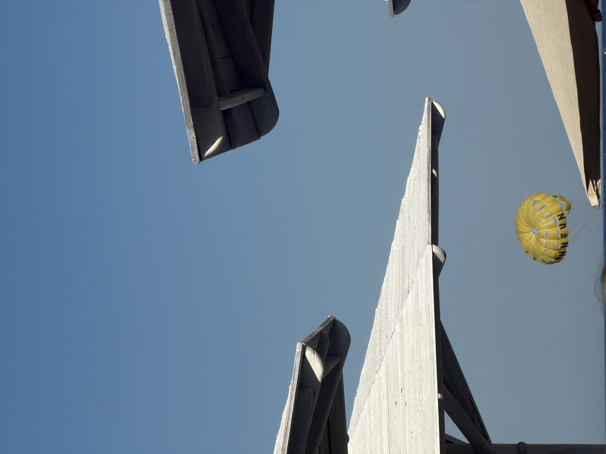 This image captures a parasail, yellow with black lettering that reads "FUN", suspended in a clear blue sky. Three white beach umbrellas with wooden frames are visible, positioned at different angles and heights, with the sky forming the background behind them. The umbrellas appear to be a part of a resort or beach area. The scene is likely located in Sharm El Sheikh, Egypt, a popular tourist destination known for its beaches and water sports.