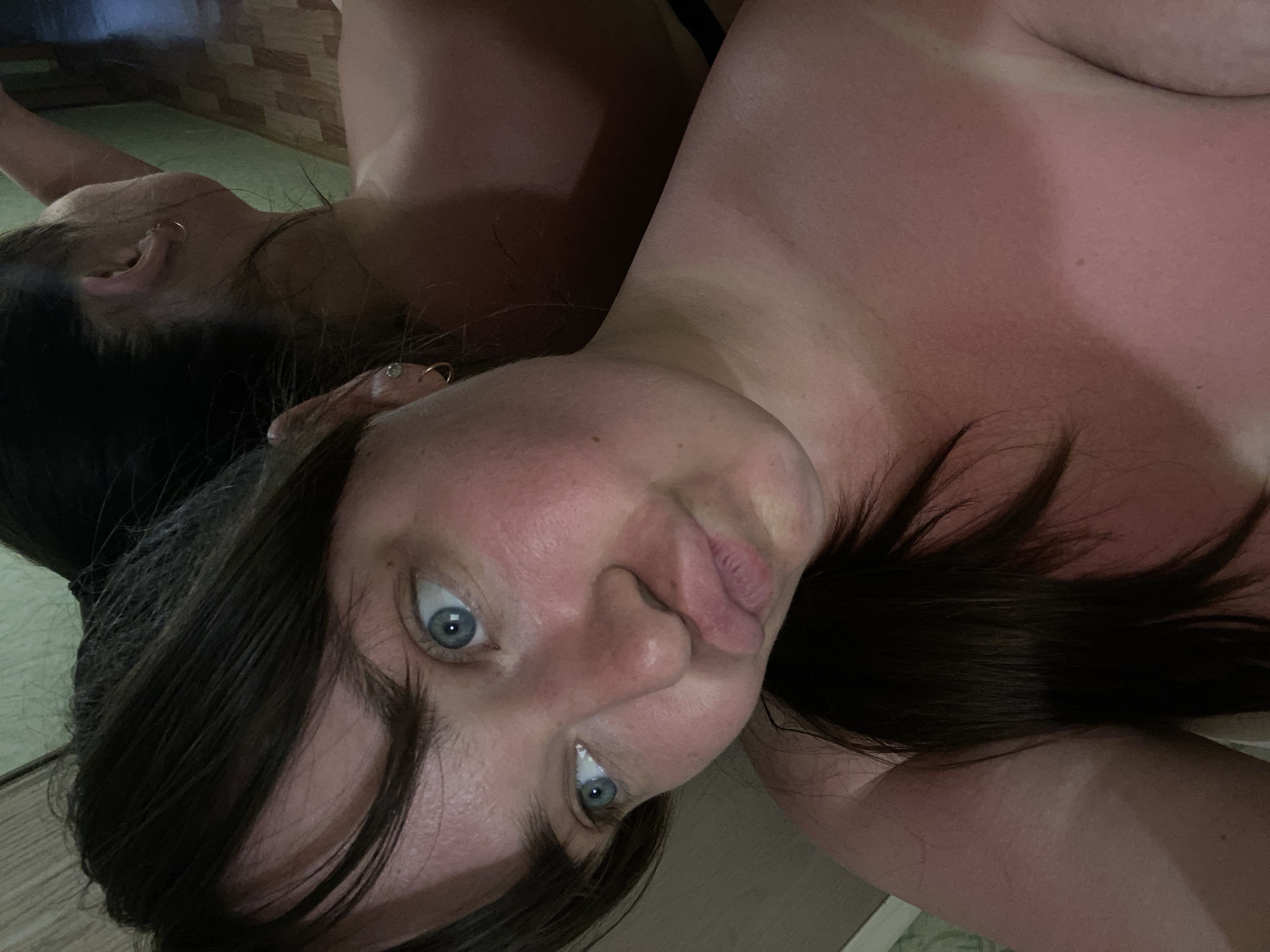 A close-up, tilted selfie of a woman with fair skin and dark hair. She has blue eyes and is looking directly at the camera. Her skin appears slightly red, possibly due to sun exposure. Part of her reflection can be seen in the upper left of the image. The background is indistinct, showing a wall with a patterned surface. There is no indication of the location being Votkinsk, Russia.