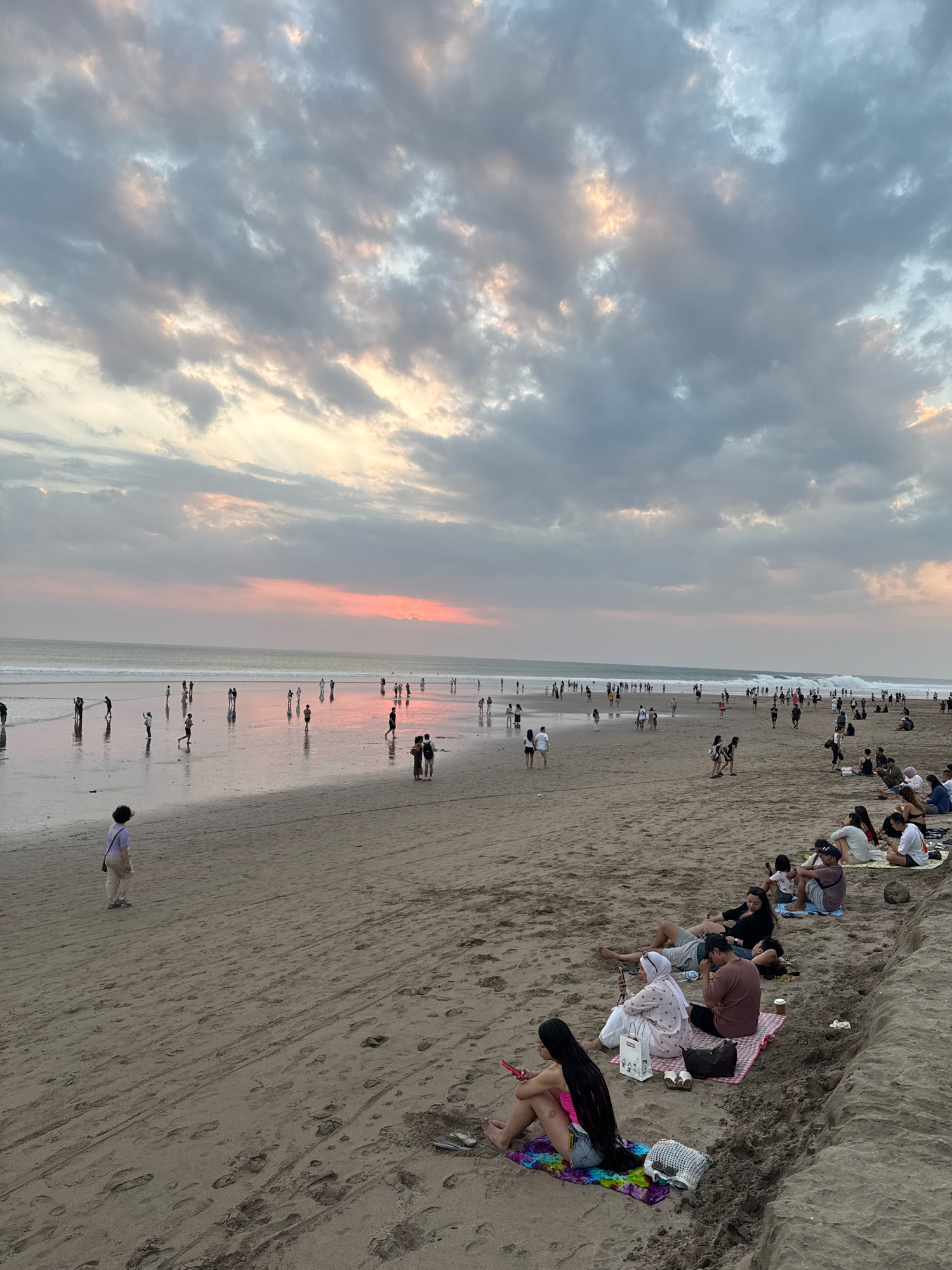 The image depicts a wide, sandy beach at sunset in Kuta, Indonesia. The sky is filled with dramatic clouds, tinged with hues of pink and orange from the setting sun, which is low on the horizon and visible between the clouds. The ocean has moderate waves crashing onto the shore, and the water reflects the colors of the sky.
There are numerous people scattered across the beach. In the foreground and mid-ground, groups of people are seated or lying on towels and blankets, relaxing on the sand. Some individuals are walking along the shoreline or near the water's edge, while others are standing or in small groups. A lone person in a white shirt and light-colored pants is walking on the sand in the left mid-ground. Further in the distance, a larger crowd of people are visible along the water's edge and in the shallow water, many appearing to be walking or standing.
The sand is a light brown color and shows numerous footprints. The overall scene is one of a busy public beach enjoying the evening twilight.