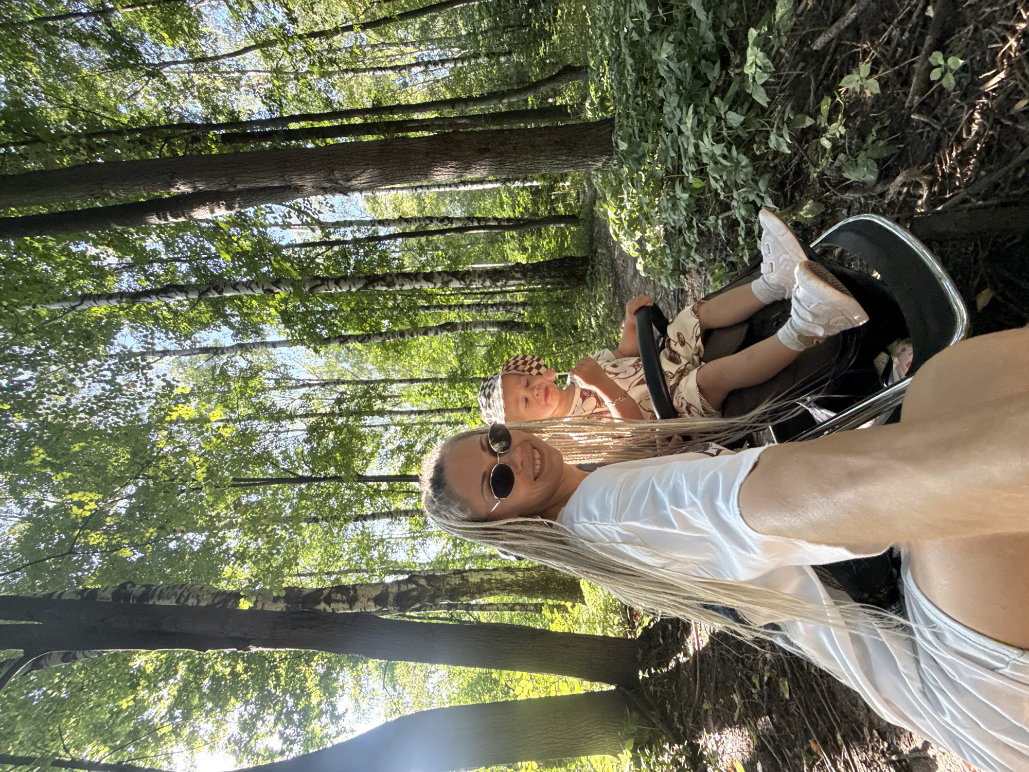 An outdoor, high-angle, medium shot captures a woman with blonde dreadlocks and sunglasses, wearing a white top, smiling at the camera. She is in a stroller next to a young child wearing a patterned hat and light-colored clothing, also seated in the stroller. The stroller is positioned on a path in a forest, with tall trees and lush green foliage surrounding them. The lighting suggests a sunny day, with sunlight filtering through the leaves. The scene appears to be in the village of Rusanovo, Russia.