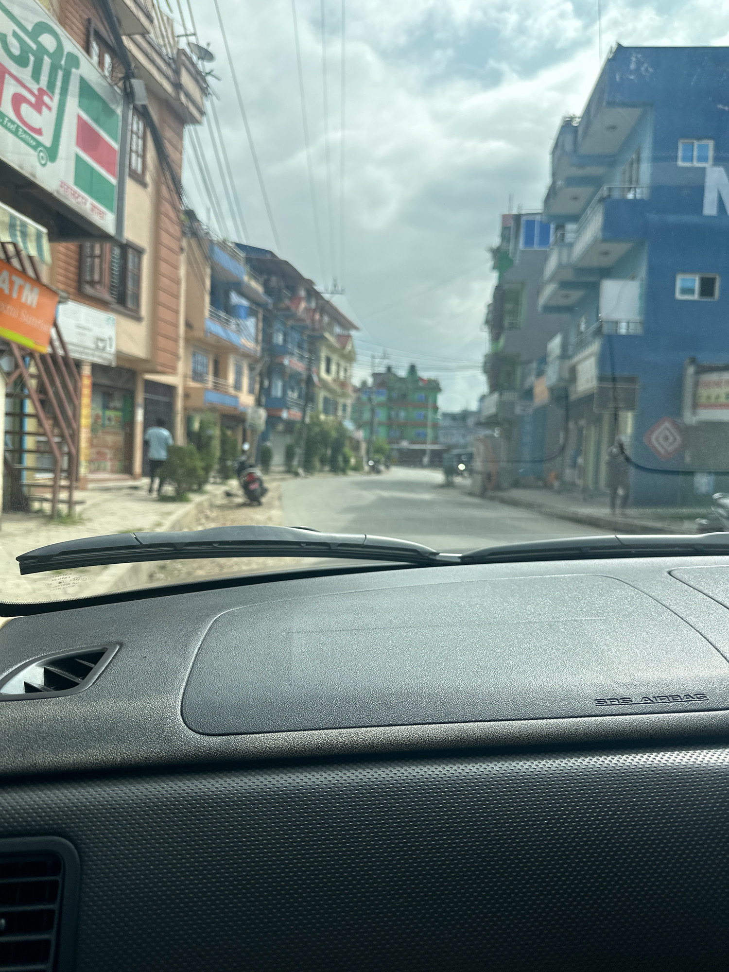 The image provides an indoor, forward-facing perspective from within a vehicle, capturing a street scene in Kageshwori Manohara, Nepal. The foreground is dominated by the vehicle's dashboard, featuring a textured black surface with a visible air vent on the left and a subtle "SRS AIRBAG" emblem on the right. The windshield, slightly grimy, offers a view of the exterior.
The street is lined with multi-story buildings on both sides, displaying a mix of architectural styles and colors, predominantly muted earthy tones and vibrant blues. Many buildings feature balconies and numerous visible electrical wires crisscross the sky. The street itself appears paved, with a slight curve guiding the view into the distance.
Several people are present: a lone individual wearing a light-colored shirt and dark pants walks away from the camera on the left side of the street, near a storefront. Another individual, obscured and partially visible on the right side of the street, appears to be walking. A red and black motorcycle is parked on the sidewalk on the left. Further down the street, traffic is suggested by indistinct shapes of vehicles, including what appears to be a small truck or van.
The environment appears to be a commercial area, with storefronts visible along the street. Signs with text, some in a non-Roman script, are mounted on the buildings. The sky is overcast with clouds, suggesting a possibly humid or rainy day. The overall impression is of a busy, lived-in urban environment.