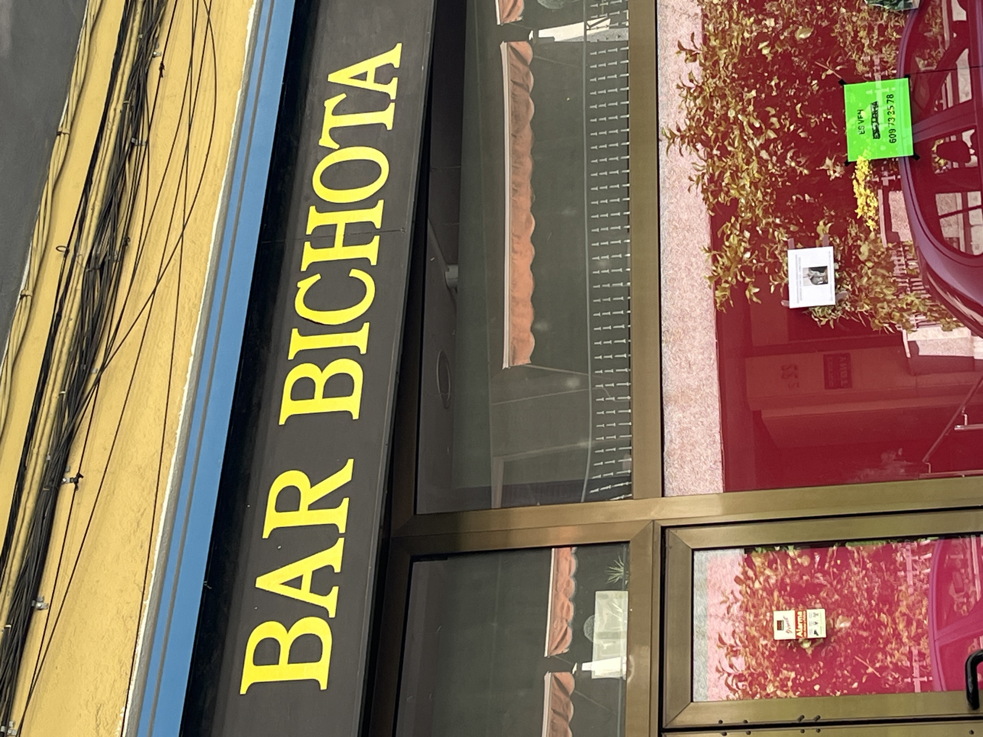 The image shows the exterior of a building with a sign that reads "BAR BICHOTA" in large yellow letters against a black background. The building has a yellow facade with black trim and a blue stripe. Below the sign, there are large glass windows with orange awnings visible inside. To the left, there is a glass door with an "Alarma" sticker and a red car reflected in the glass. To the right, there is a red wall and a green sign with text and a phone number. The context of Figueres, Spain, is mentioned but not visually evident in the provided image. The scene appears to be an outdoor view of a commercial establishment during daylight hours.