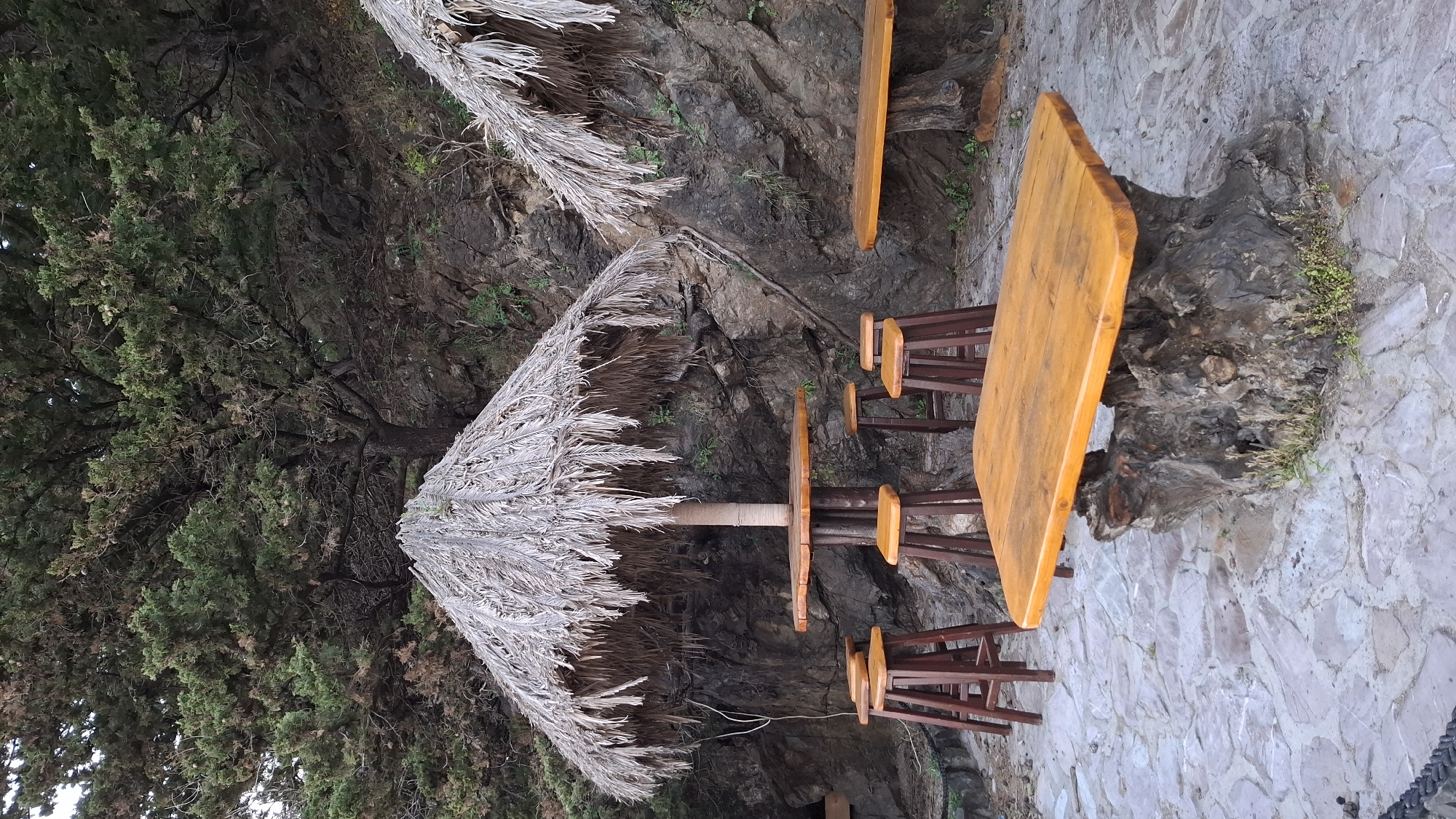 The image shows an outdoor seating area in Boreti, Montenegro. There are two wooden tables, each surrounded by three wooden stools with metal frames. A thatch umbrella provides shade for one of the tables. The area is adjacent to a stone cliff face and a large green tree. The ground is paved with irregularly shaped stone slabs.