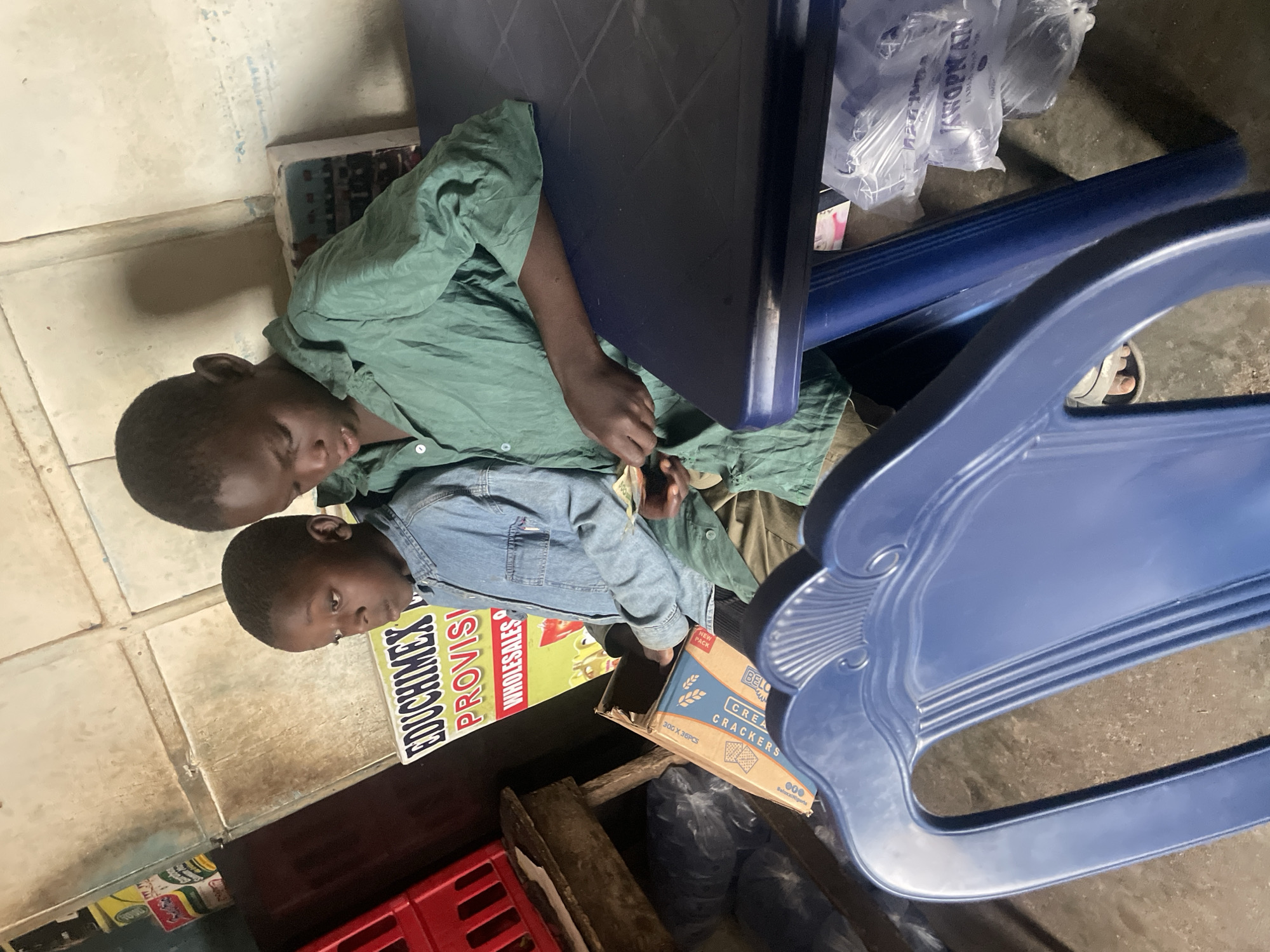 Two young African boys are lying down in an interior space. One is wearing a light green long-sleeved shirt and the other is wearing a denim jacket. They are resting on a poster advertising "EDUCHIMEX PROVISI WHOLESALES." Nearby are a box of "Bello CREA CRACKERS" and various pieces of blue plastic furniture including a table and a chair. There is a red plastic crate visible in the lower left corner. The scene is cluttered, with plastic bags and various objects scattered around. The location is presumed to be Barakin Lerawi, Nigeria, based on the OCR text found on the advertisement, and cracker box.