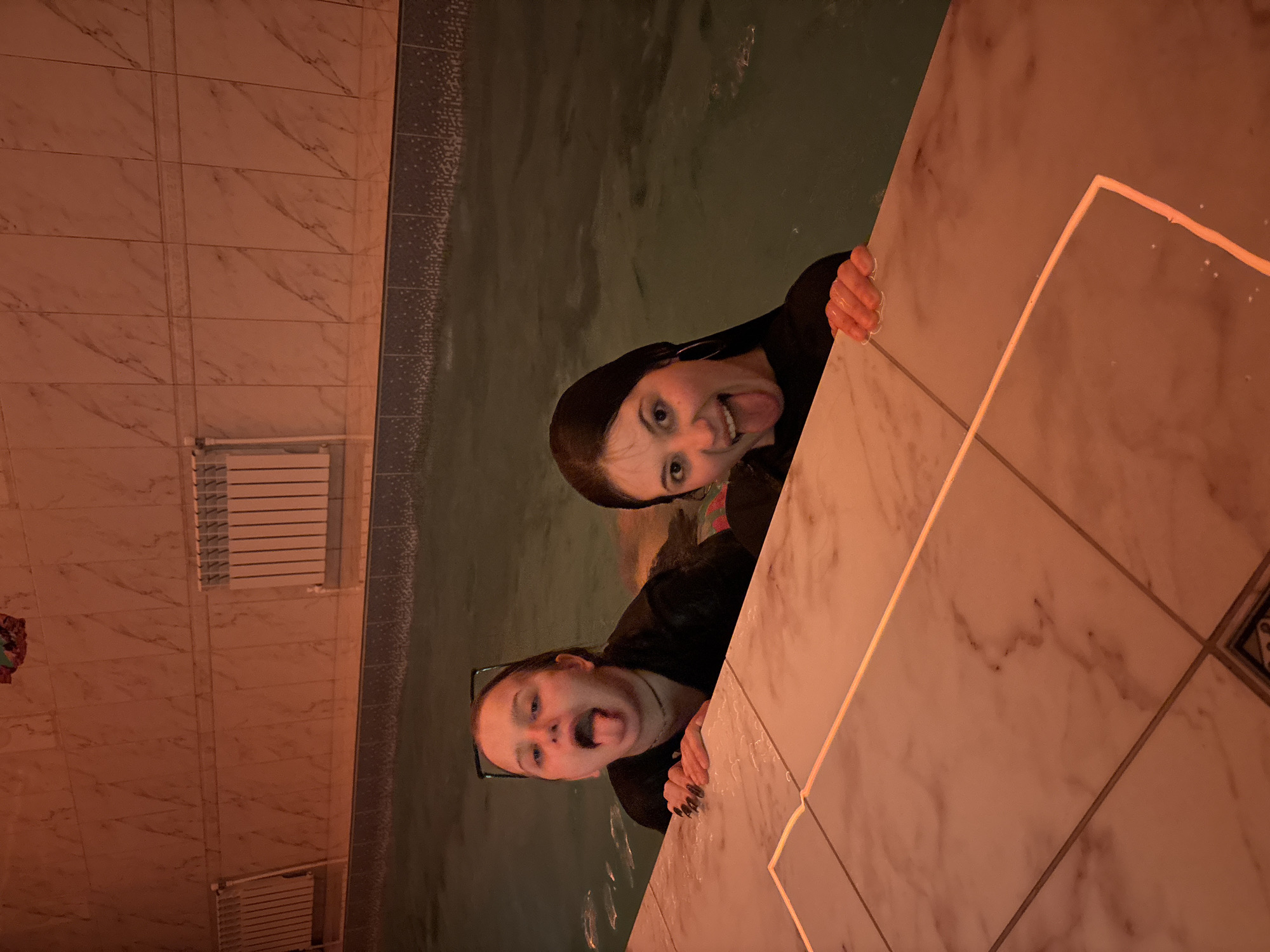 Two females are peering over the edge of a tiled pool, sticking their tongues out. The pool is surrounded by marble tiling. The location is likely a pool in Chaikovsky, Russia. Radiators are affixed to the tiled wall.