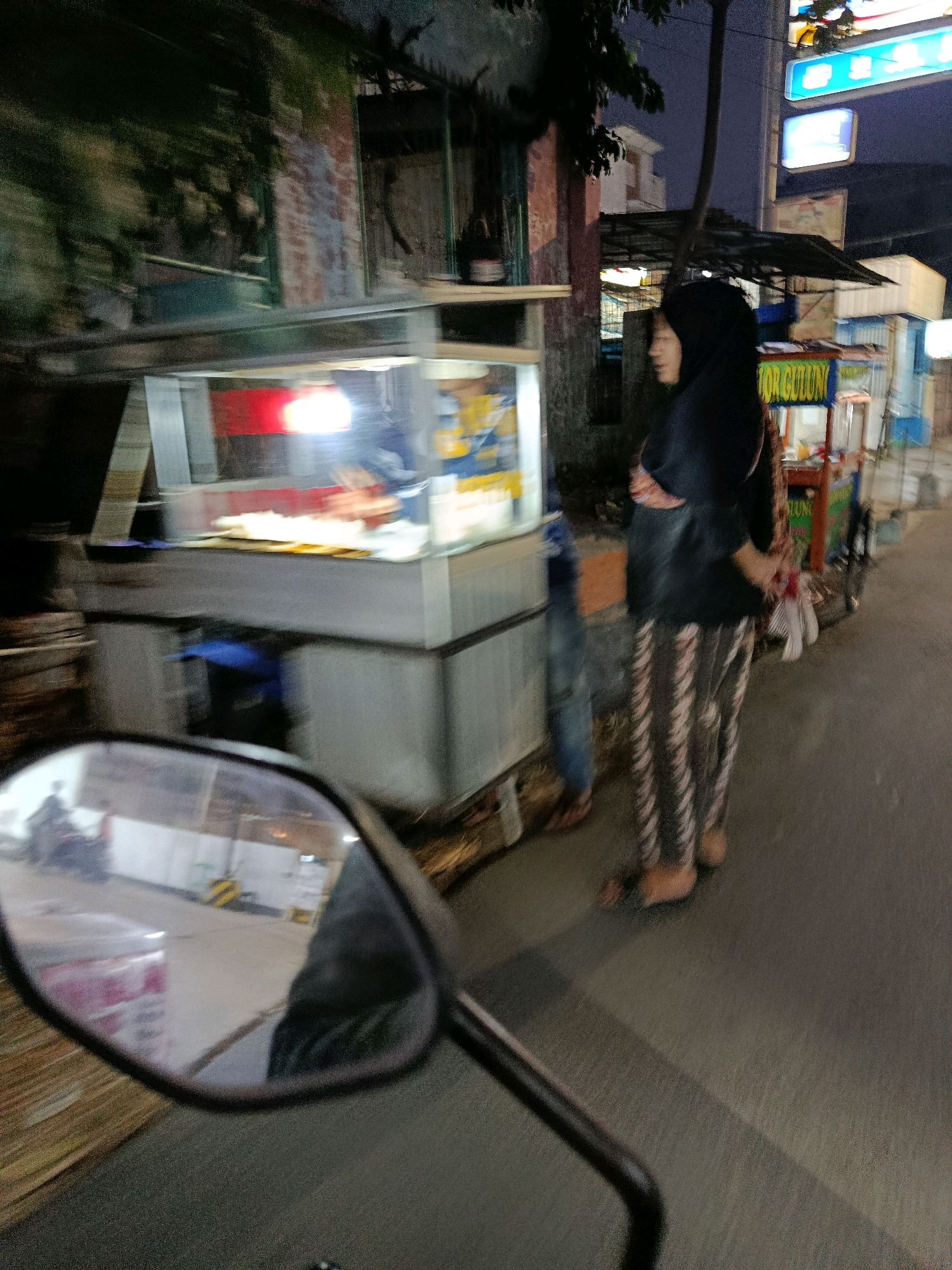 The image depicts a street scene at night in Bekasi, Indonesia. A woman wearing a black hijab and patterned leggings stands next to a food stall. The food stall has a glass display case with food items inside and is illuminated by lights. In the foreground, a motorcycle's side mirror is visible, reflecting parts of the street and possibly the rider. Other elements in the background include buildings, trees, and streetlights. The overall atmosphere suggests a bustling urban environment after dark.