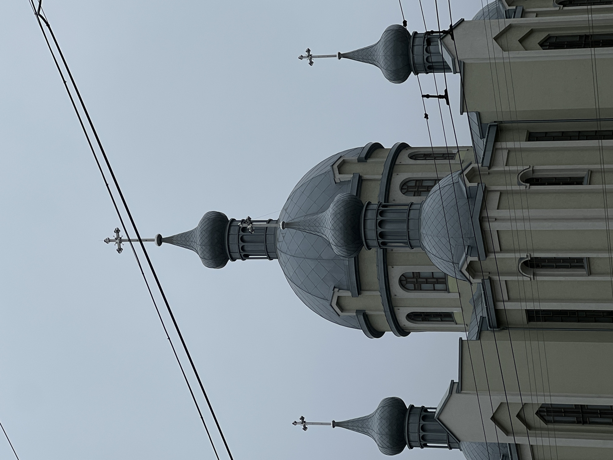 The image captures the exterior of a large religious building, likely an Orthodox church or cathedral, characterized by its distinctive architecture. The building features several prominent onion domes, predominantly gray and textured, with the largest central dome flanked by smaller ones. Each spire is topped with an Orthodox cross. The main structure is a light, possibly pale green or beige color, with visible windows of both arched and rectangular designs.
Several dark utility lines or power cables are stretched diagonally across the upper and left portions of the frame, obscuring part of the clear, overcast sky that forms the background. The lighting suggests a cloudy daytime scene. There are no people visible, and no specific activity is taking place. The image is oriented sideways, appearing rotated 90 degrees clockwise. This architectural style is typical of Eastern Europe, and the provided context places the location in Ternopil, Ukraine. No visible text is present.