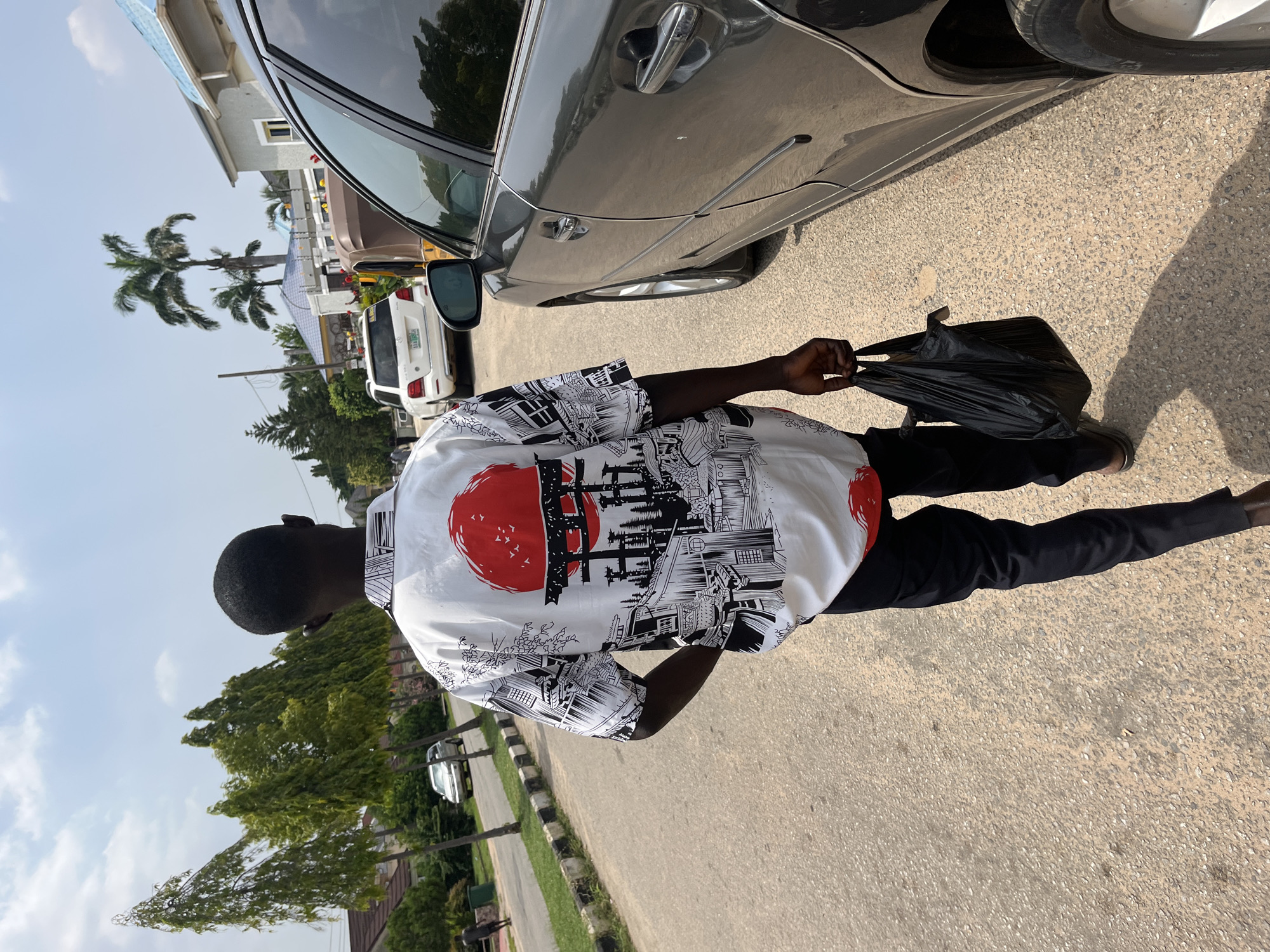 A person walks down a paved street in Abuja, Nigeria, carrying a black plastic bag in their right hand. They are wearing a white short-sleeved shirt with a black and red Japanese-inspired print and black trousers. The person is walking away from the camera, with their back to the viewer. To the right of the person, a dark grey car is parked on the street. The street is lined with trees on the left side, and residential buildings with blue and grey roofs are visible in the background. The sky is clear and blue with some scattered clouds.