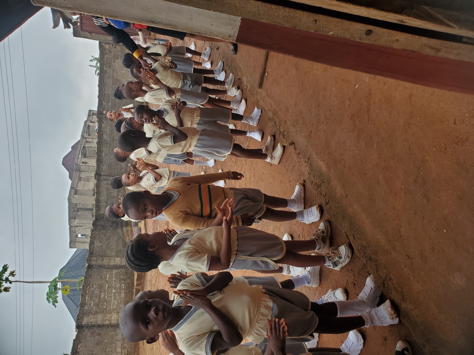 A medium shot, taken at eye level, captures a line of approximately 15 African children, facing the same direction, in an outdoor setting. The location is identified as Ukwu-Abwa, Nigeria.
The children are dressed in school uniforms consisting of a short-sleeved, light-colored shirt or sweater, grey shorts or skirts, white socks, and sneakers. They are positioned in a line on a patch of reddish-brown dirt. Their hands are clasped together, appearing as though they are clapping or praying.
In the background, there is a light grey brick building, part of which is under construction. Additionally, there are trees and vegetation. Overhead, the sky is overcast and grey. A portion of a building and a shadowed corner are visible at the right corner of the frame.