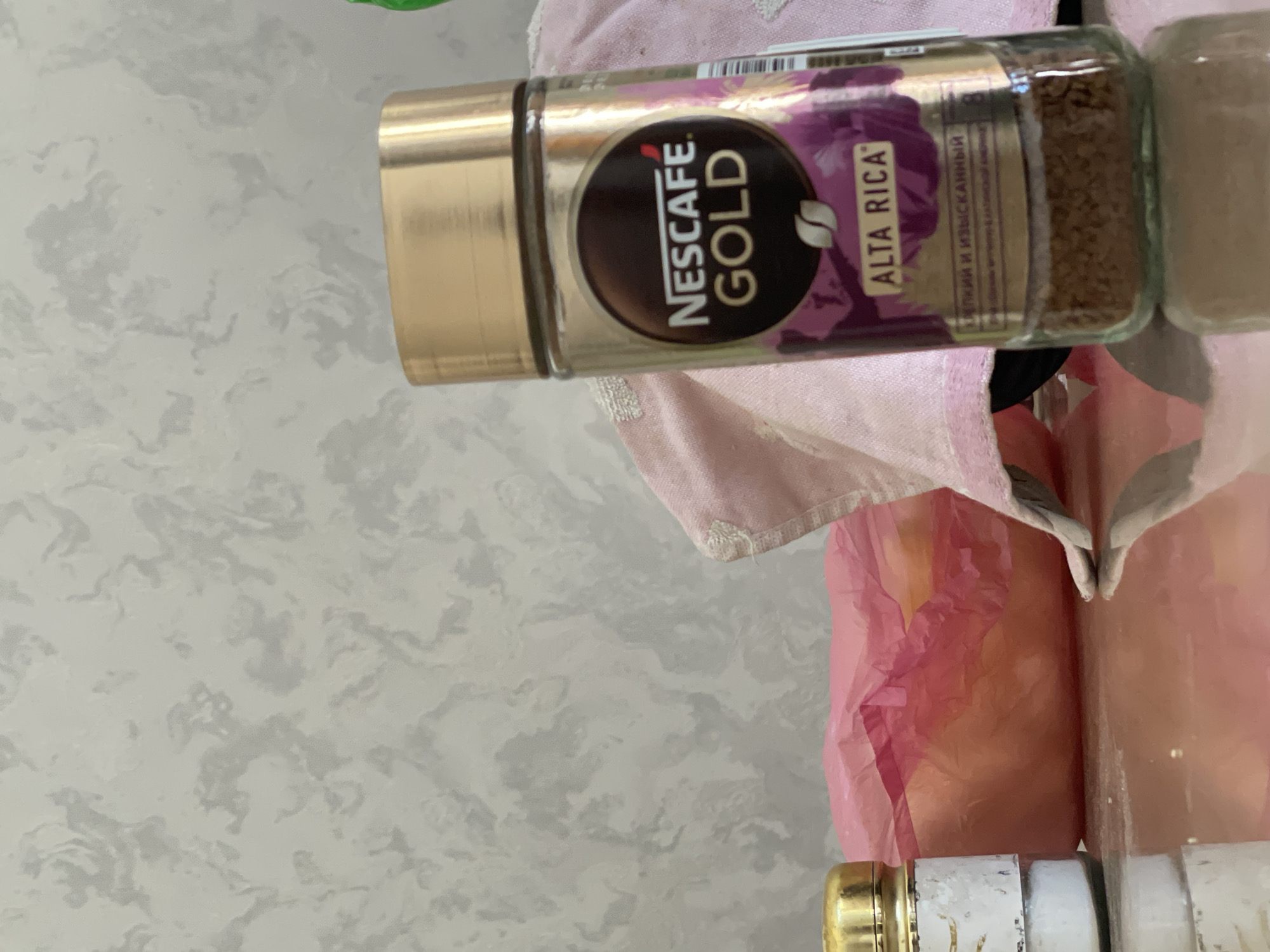 The image displays a close-up of a glass jar of Nescafé Gold Alta Rica instant coffee. The jar has a gold lid and a label featuring the Nescafé Gold logo and floral imagery. The coffee grounds are visible at the bottom of the jar. To the left of the coffee jar, there is a smaller white and gold container, likely a spice jar or salt shaker. Behind the coffee jar, a pink fabric or cloth is partially visible. The items are placed on a reflective surface, possibly a table. The background consists of a textured gray wall. The scene appears to be indoors, in a domestic setting. There is no indication of the specific location within Tashkent, Uzbekistan.