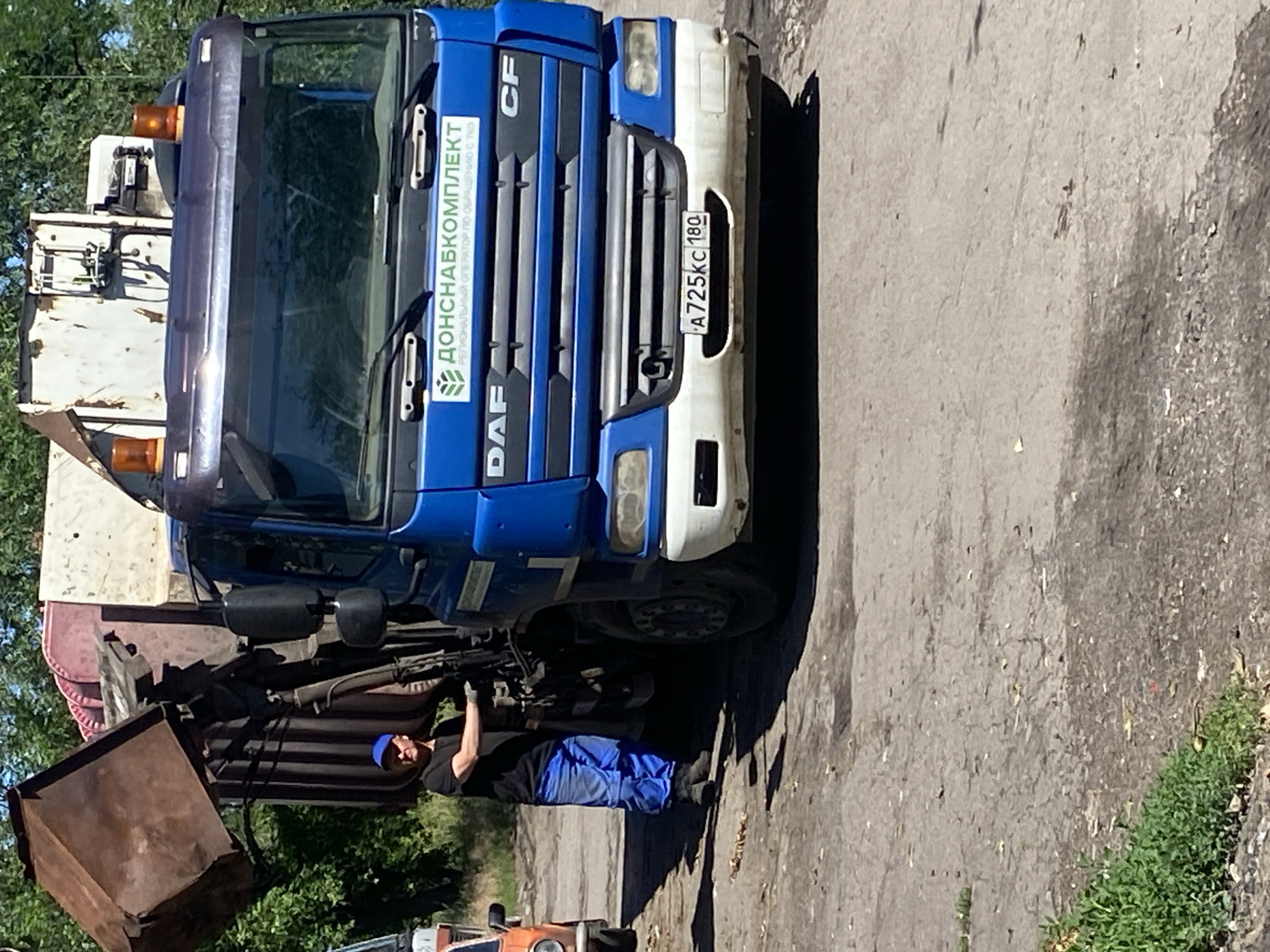 A man in a blue cap, black t-shirt, and blue overalls is standing next to a blue DAF garbage truck. The man is operating the hydraulic arm of the truck, which is lifting a large, rusty bin. The truck has a logo with Russian text and the license plate "A725KC 180". The scene appears to be on a paved area with some greenery and trees in the background. The sky is clear and the lighting suggests it is daytime.
