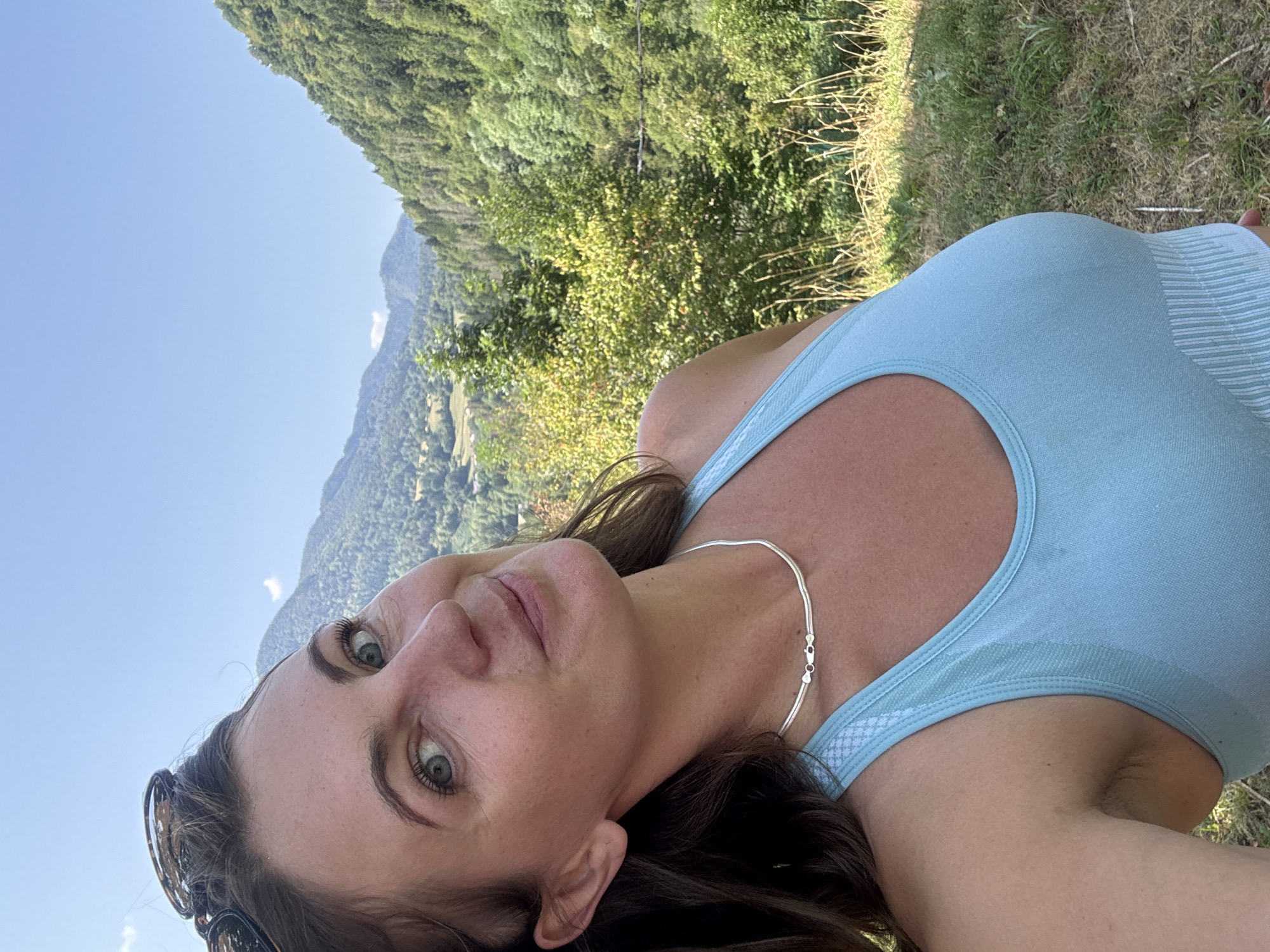 A close-up selfie shows a woman in a light blue athletic top and a silver necklace. She has fair skin, green eyes, and dark brown hair. She is wearing sunglasses on her head. The background features a lush green mountain landscape with trees under a clear blue sky. The image appears to be taken outdoors, possibly in the countryside, but there is no direct evidence to confirm it is in Saint-Claude, France.