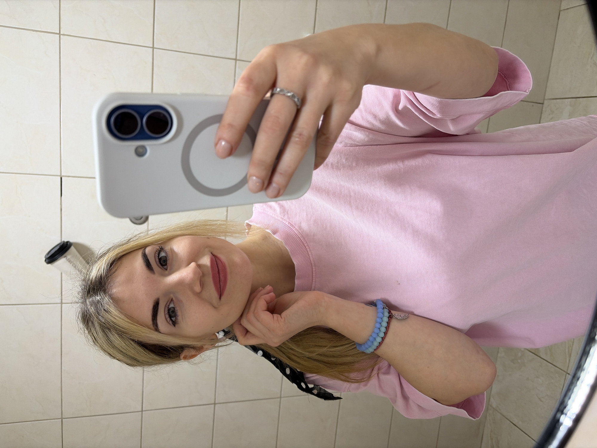 A female subject is depicted taking a mirror selfie indoors. She has blonde hair, parted centrally, with a black and white polka dot scrunchie visible on the left side of her head. Her facial features include light makeup with red lipstick, and a slight smile. She wears a light pink short-sleeved t-shirt. Her left wrist displays two beaded bracelets, one light blue and one red, with a small silver charm. Her right hand, holding a light grey/white smartphone, exhibits a patterned silver ring on the ring finger. The smartphone features a dual-lens camera module and a prominent circular magnetic attachment ring on its rear case. The subject's left hand is positioned near her neck and ear. The background consists of light beige square tiles with dark grout lines. A white disposable coffee cup with a black lid is visible leaning against the tiled wall in the upper right portion of the image. The specific location within Budva, Montenegro, cannot be determined from the visual evidence, though the tiled environment is consistent with a public or private restroom.