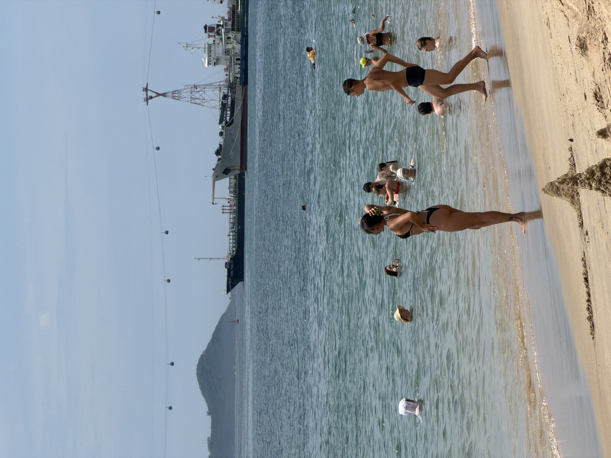 The image depicts a sunny day at a beach in Nha Trang, Vietnam. Several people are enjoying the water, with some wading and others swimming. In the background, a large cargo ship is docked near a tower with cables extending across the water, suggesting a cable car system is in operation. A small, sandy hill is visible in the foreground on the beach. The sea is calm, and the sky is clear with a few scattered clouds.