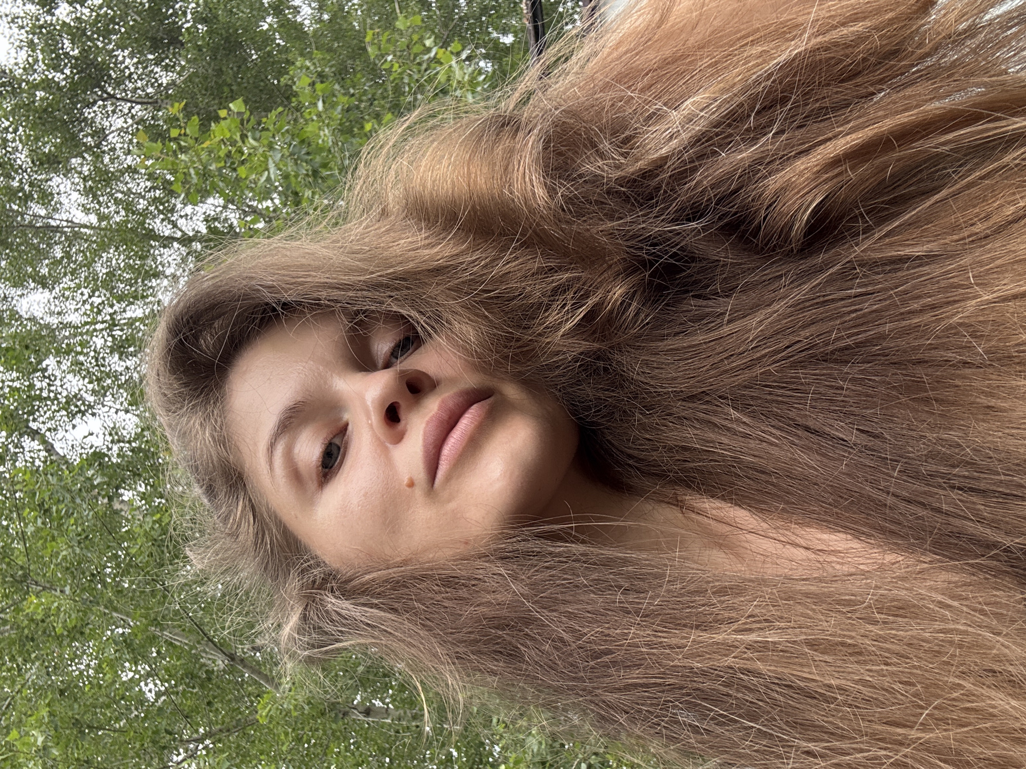 The media file is a close-up, low-angle selfie of a young woman outdoors. She has long, light brown hair that frames her face, with some strands falling forward. Her face is tilted upwards, and her eyes are looking towards the camera. She has fair skin, with a small mole on her cheek. Her lips are slightly parted, revealing a natural, soft pink color.
The background consists of lush green foliage, suggesting a natural setting with trees. The lighting indicates it is daytime, likely overcast or in shade, as there are no harsh shadows. The overall mood of the image appears relaxed and natural, with the woman's gaze and expression conveying a sense of calm. There is no visible text in the image. The location context is Minusinsk, Russia.