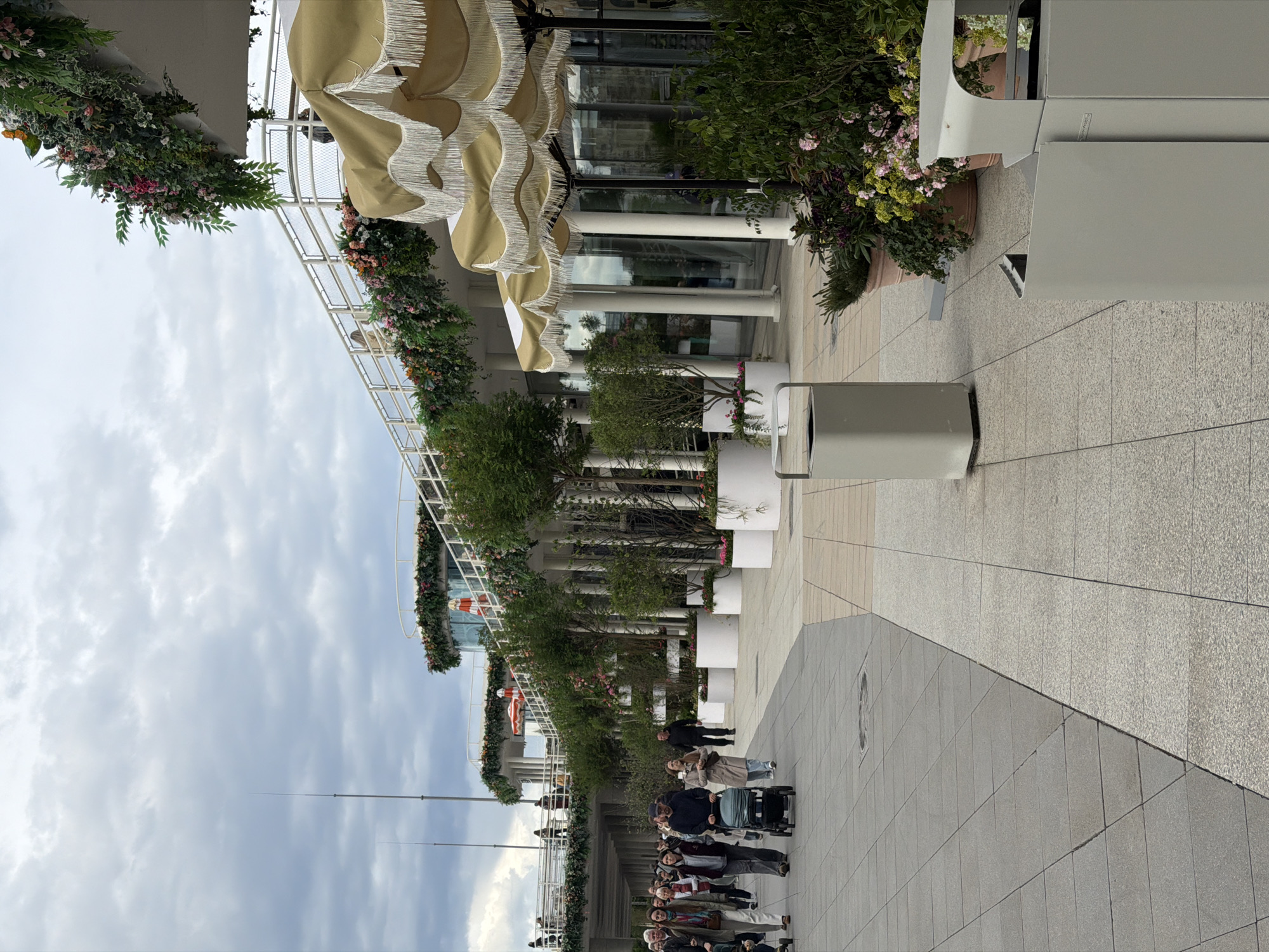 The image shows a street in Moscow, Russia, featuring a building decorated with greenery and awnings. A group of people is walking along the sidewalk. There are trash cans and potted plants along the street. The sky is cloudy.