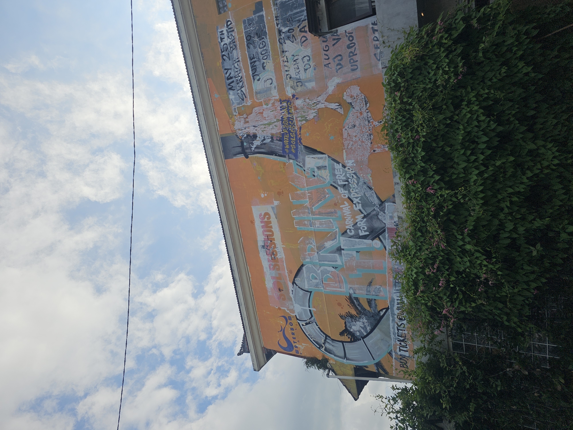 The image features a tall building in North Kuta, Indonesia, adorned with a large, colorful mural. The mural is painted in orange and gray tones, displaying various abstract images, including a stylized palm tree and the text "SOLSESSIONS" and "CARNIVAL OF FREE FORM EXPRESSION". Also visible are faded dates and names such as "MAY 25TH SKRATCH BASTID," "JUNE 29," "JEREMY SOLE," "DJ DAY," and "DJ VA UPROOT." Dense, green vines heavily cover the lower right portion of the building's facade, partially obscuring the mural and a wire mesh fence. In the background, a blue sky with scattered white clouds can be seen. A lone power line stretches across the sky to the upper left.
