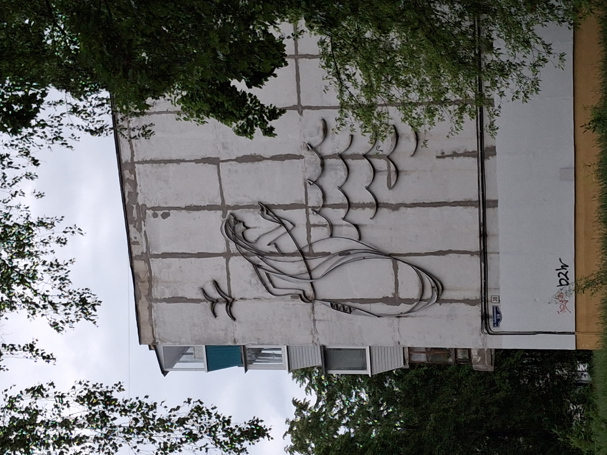 The image shows a building in Chaikovsky, Russia. A metal sculpture of a fish is mounted on the side of the building. The building is made of white concrete panels, and the metal sculpture is made of smooth, flat metal. Graffiti is visible near the bottom of the building. Trees obscure the view. Windows and building utility infrastructure are visible at the base of the building. The street address is visible as "59" on a blue sign.