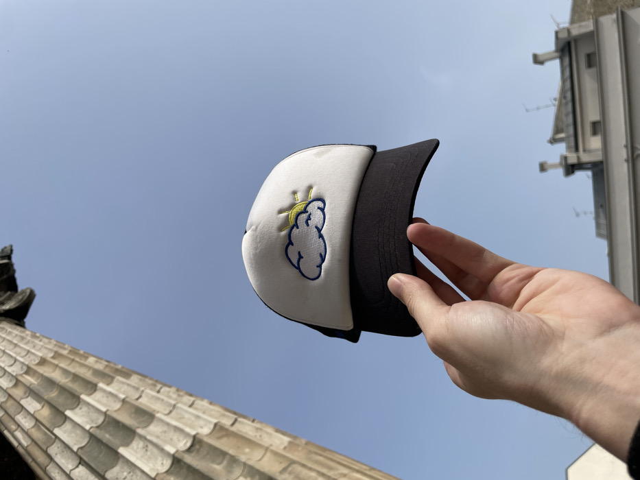 A human right hand is prominently displayed in the lower center, holding a black and white trucker hat by its brim. The hat features a white front panel with an embroidered design of a blue cloud partially obscuring a yellow sun. The hand and hat are positioned against a clear, bright blue sky. To the upper left, a large, light-colored classical column, exhibiting fluting and an ornate capital consistent with Corinthian or Composite architectural orders, extends diagonally from the top edge. In the lower right background, a cluster of distant Parisian Haussmannian-style rooftops with multiple chimneys and antennas is visible. The scene is located in Paris, France, with the architectural elements suggesting proximity to a major landmark such as the Panthéon or La Madeleine, viewed on a sunny day.
