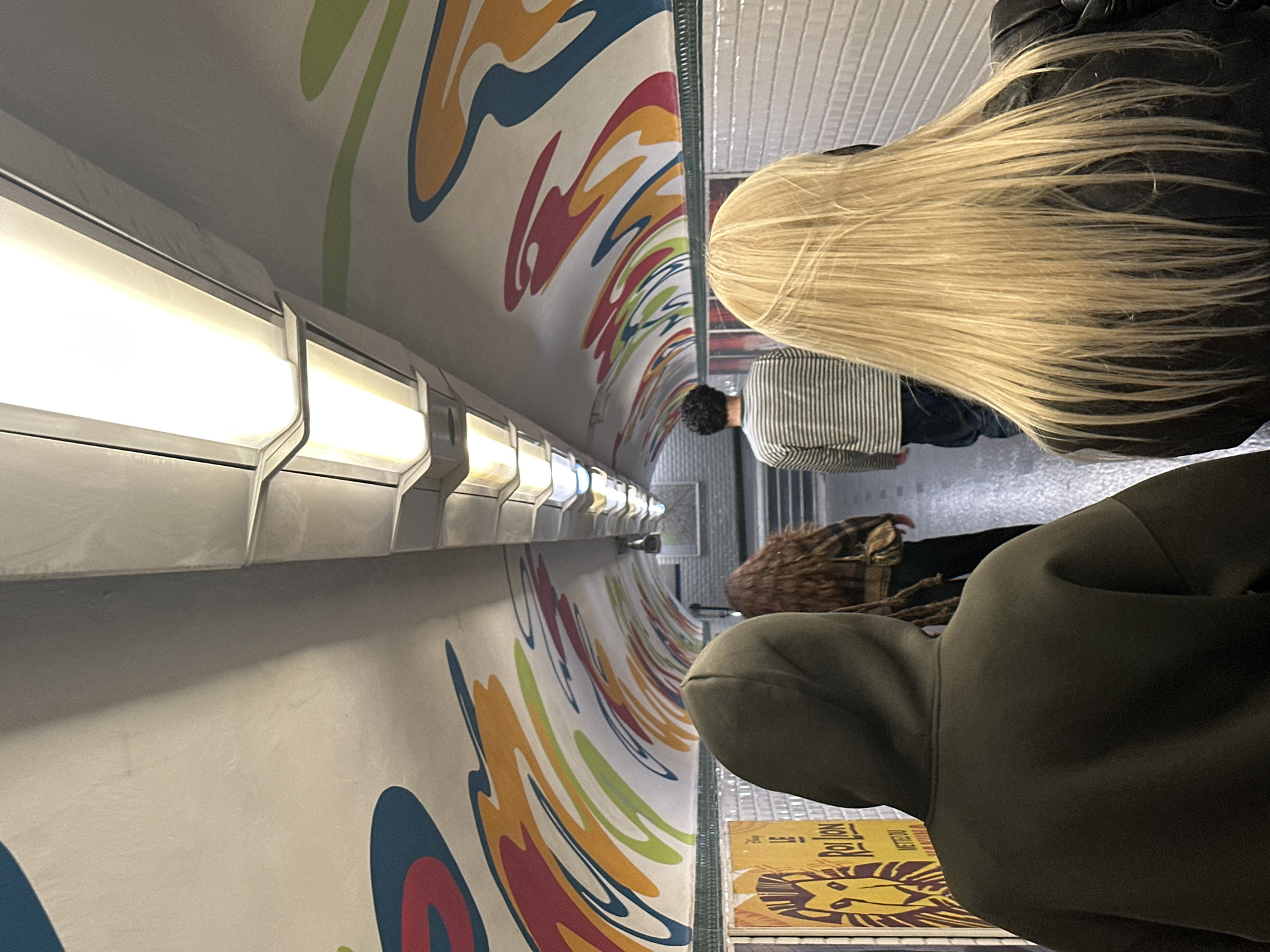 The image shows the interior of a Paris Métro car, specifically decorated with a colorful mural in shades of blue, yellow, red, and green. An overhead light fixture runs the length of the car's ceiling. Three individuals are partially visible in the foreground. The first has long, blonde hair and is wearing a black jacket. Another individual has brown hair and is further in the distance. A third individual is wearing a green hoodie. A poster for "Le Roi Lion" ("The Lion King") is visible at the bottom of the frame. The walls of the Métro car are lined with white subway tiles. The scene appears to be taking place on a Paris Métro line that has been artistically decorated.