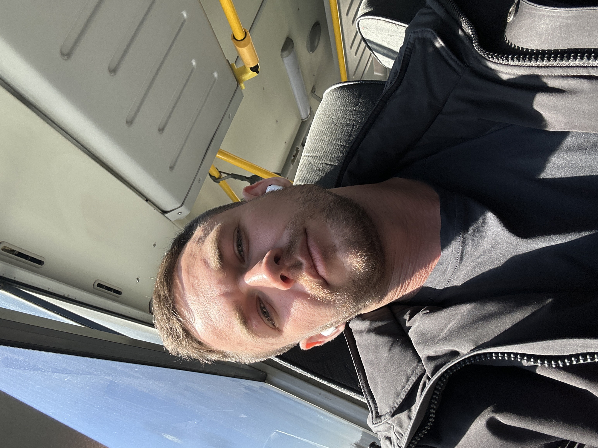 A man is sitting on a bus, possibly in Perm, Russia. He is fair-skinned, with short brown hair and a beard. He wears a black jacket and a black t-shirt. He has a white earbud in his ear. The bus has yellow handrails and a grey interior. Outside the window, the sky is clear and blue. The image appears to have been taken from a low angle.