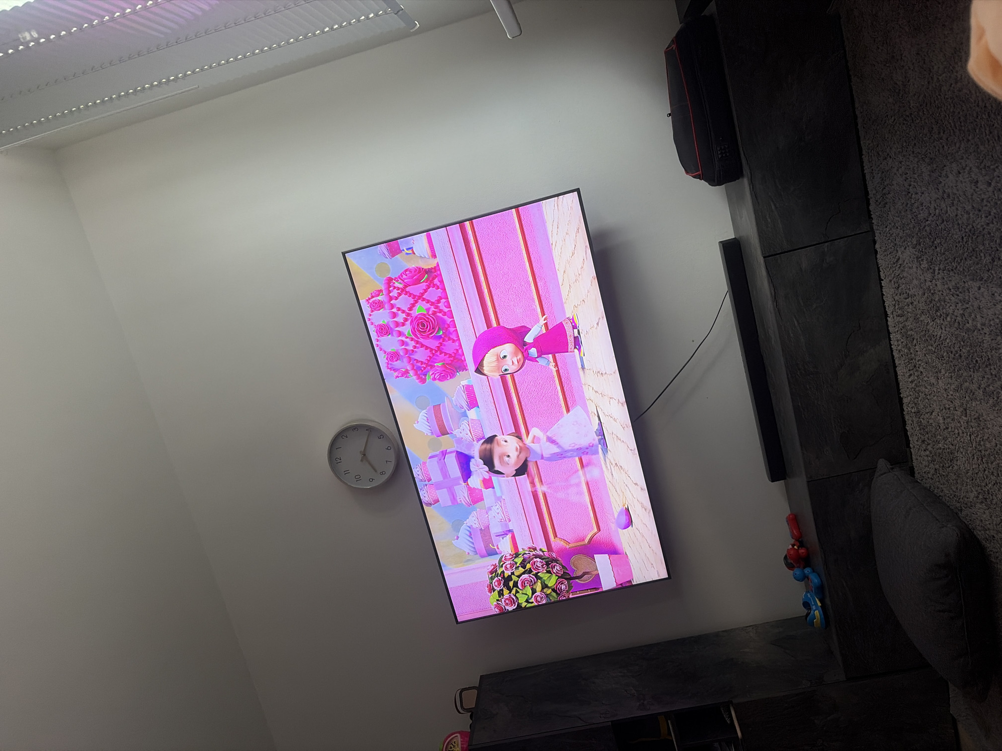 The image depicts a living room interior, specifically a view towards a wall with a television. The television screen displays an animated scene featuring two young female cartoon characters in a pink-themed setting with cupcakes and floral decorations. Above the television, a round analog clock is mounted on the wall, showing the time as approximately 10:08. To the left of the television, a dark gray tall cabinet with shelves is visible. In front of the television, a dark gray media console is present, with a soundbar on top and some toys on its surface, including a blue and yellow toy car and a red toy. A large gray floor cushion is in the foreground, resting on a textured gray carpet. To the right of the television, a dark bag with red trim is placed on the media console, and a window with white blinds is partially visible. The overall scene suggests a domestic setting, likely a living room where entertainment is being viewed. The image does not contain any explicit or implicit information that would allow for the determination of its location within Duisburg, Germany.