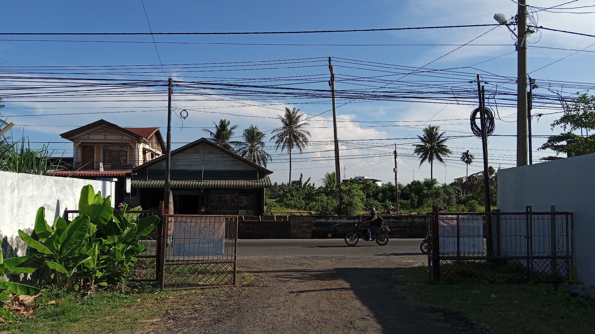 The image depicts a street scene in Bireuen, Indonesia, likely during the daytime given the bright sunlight and blue sky with scattered clouds. In the foreground, a dirt path leads towards a paved road. To the left, a large cluster of banana plants with broad, green leaves is visible behind a metal gate. A white wall borders the left side of the scene.
Across the road, there are several structures. A two-story house with an orange roof and decorative yellow trim is prominent. Next to it is a single-story building with a dark green awning and what appears to be a storefront. Behind these buildings, lush green vegetation, including several palm trees, covers the landscape, suggesting a tropical environment.
Several utility poles with a dense network of black wires crisscross the sky above the scene. On the road, a person is riding a dark-colored motorcycle. Another motorcycle is parked behind a metal fence on the right side of the image, partially obscured by a light blue wall. The overall impression is one of a rural or semi-urban setting in a tropical climate.