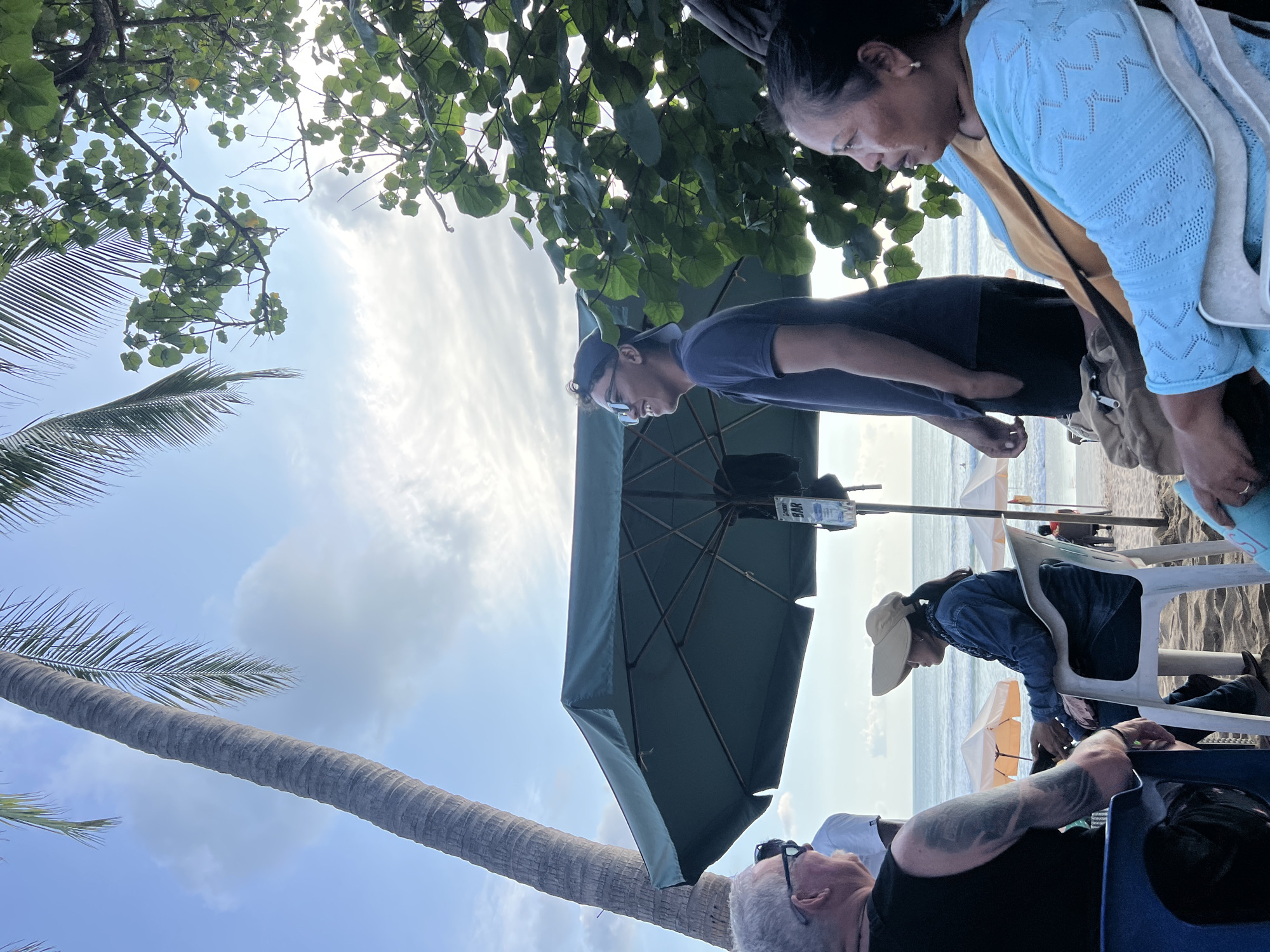 The image captures a scene on a beach in Kuta, Indonesia. In the foreground, several people are seated under a large, dark green umbrella. To the left, a man with grey hair and sunglasses is visible from the back, his tattooed arm resting on his leg. Next to him, a woman wearing a large, light-colored hat and a denim jacket is seated. To the right, a woman in a light blue cardigan over a yellow top is looking to the right. Behind her, another person's arm is visible. A young man wearing sunglasses and a baseball cap is standing and smiling, facing left. In the background, the ocean is visible with gentle waves and a few scattered beach umbrellas. Palm trees and lush green foliage frame the scene against a partly cloudy sky. A sign for "CHERRY BAR" is attached to a pole near the umbrella.