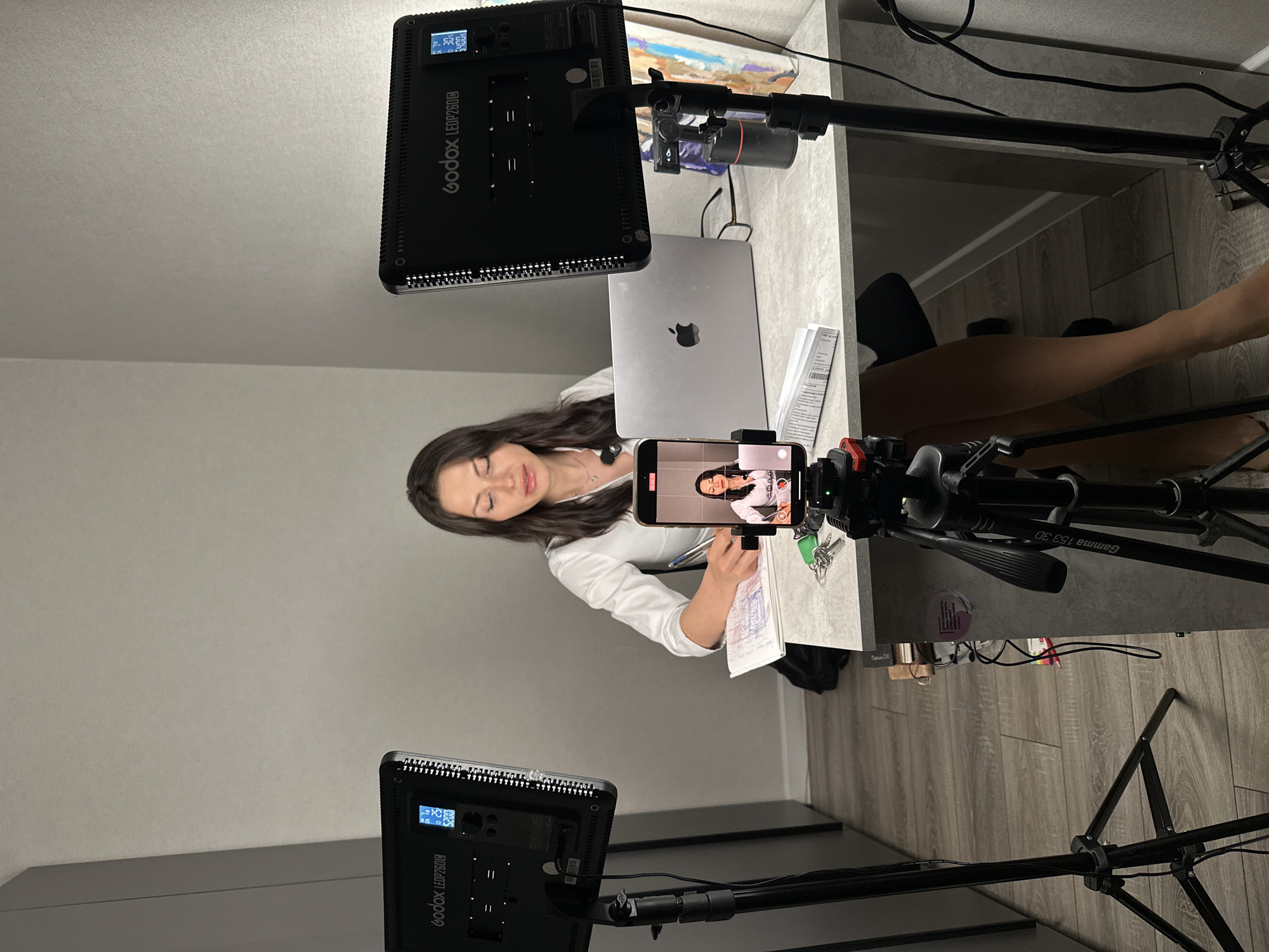 The image depicts a woman sitting at a desk in a room, possibly a home office or studio. She is wearing a white shirt and appears to be engaged in writing or studying, with papers and a laptop in front of her. A smartphone is propped up on a tripod, recording her activity, and in its screen, a smaller version of the woman is visible, suggesting she is live-streaming or recording a video. Two large LED lights, branded "Godox LEDP260C," are positioned on stands on either side of the desk, illuminating the scene. A second tripod, possibly for a larger camera, is visible in the foreground. The room has light-colored walls and a wooden floor. There are no obvious indicators of the specific city or country, but given the prompt's instruction for "Unknown, Russia," this location is assumed.