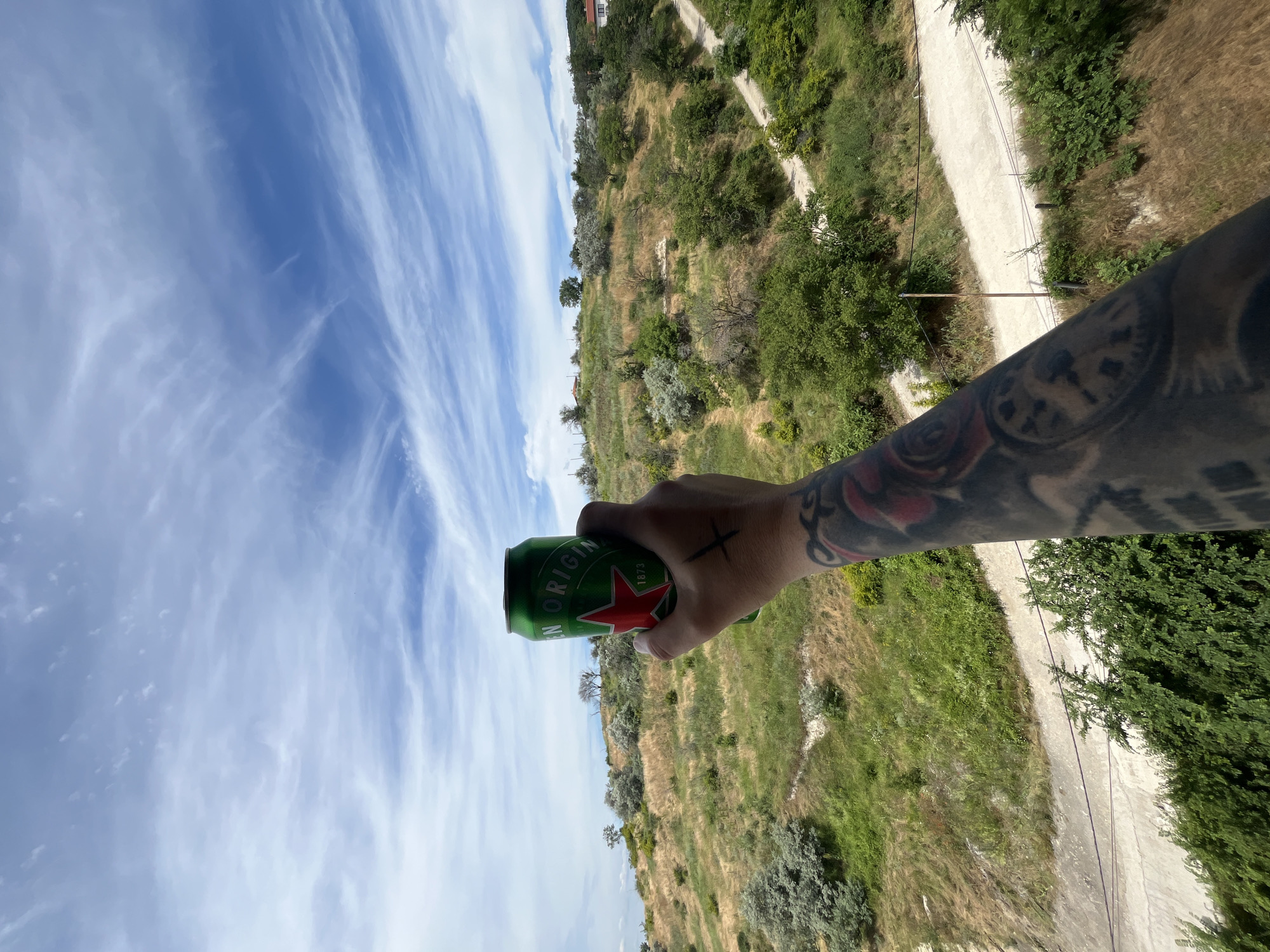 This media file captures a person's hand holding a green can of Heineken beer against a backdrop of a hilly, green landscape under a bright blue sky with wispy white clouds. The can is prominently displayed in the foreground, with the red star and the words "ORIGIN" and "1873" visible. The hand holding the can has tattoos, including a red rose and a cross symbol drawn on the back of the hand.
The scene appears to be outdoors, possibly on a sunny day, given the bright lighting. The landscape is characterized by lush green vegetation, dry grass, and a dirt path winding through the hills. In the distance, a building with a white roof and red tiles is visible on the top of one of the hills. The orientation of the image suggests it was taken from a high vantage point, looking down on the landscape.
The overall mood of the image seems relaxed and perhaps celebratory, with the act of holding a beer in a scenic setting often associated with leisure or enjoyment. The presence of the can suggests that the person is likely enjoying a drink outdoors.
The location context provided is Varna, Bulgaria, which aligns with the Mediterranean-like vegetation and the hilly terrain depicted. The time of day appears to be daytime, with clear skies and good visibility.