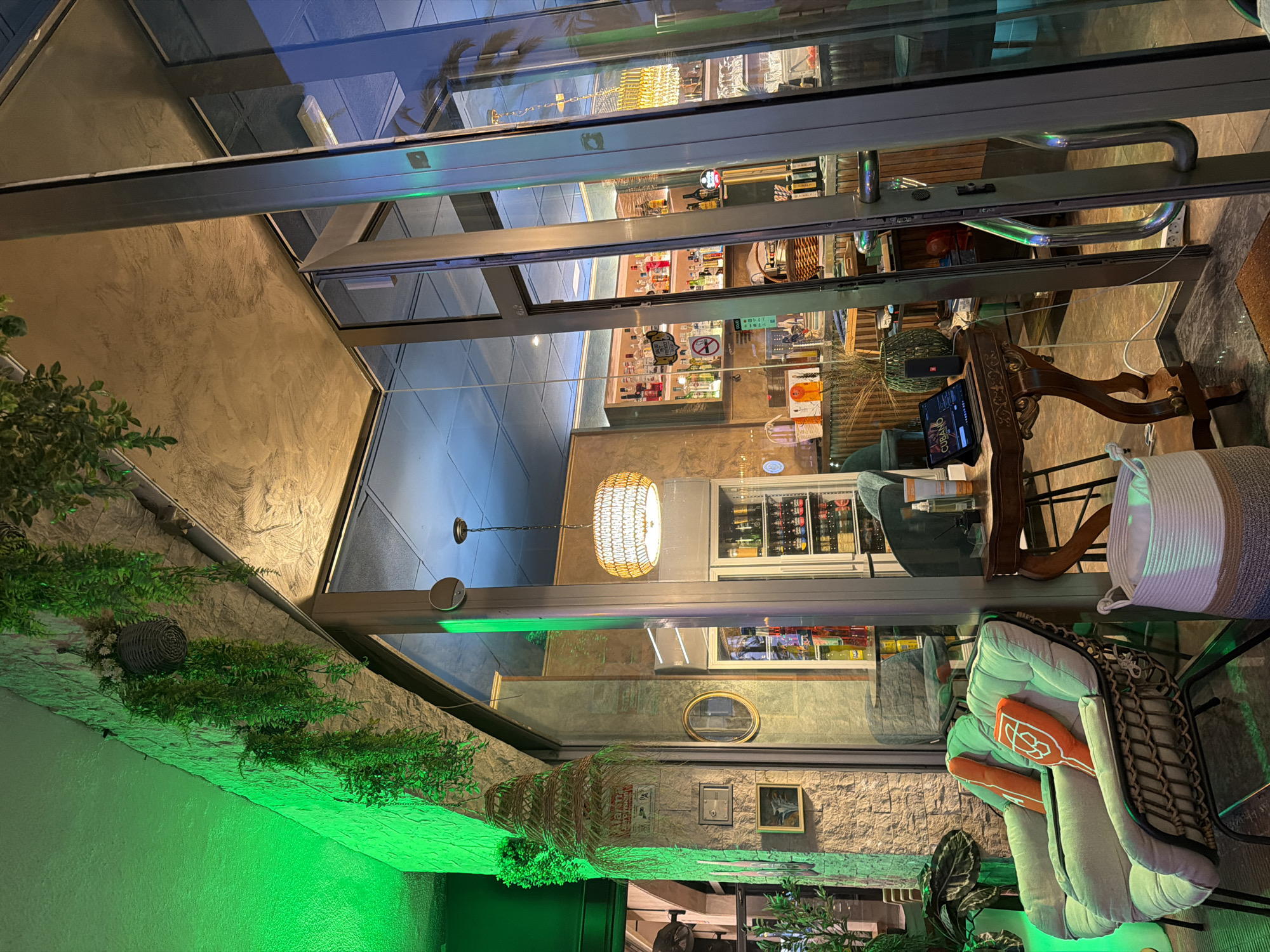 The image depicts the exterior of a modern establishment, possibly a bar or restaurant, in Salou, Spain. The scene is illuminated by vibrant green lighting emanating from the left side of the frame, casting a striking glow on a textured stone wall and lush hanging plants. The entrance features large glass doors and windows, offering a view into the interior.
Inside, a well-stocked bar with various bottles of alcohol is visible, along with a refrigerated display showcasing beverages. Contemporary lighting fixtures, including a woven pendant lamp, add to the ambiance. In the foreground, two plush armchairs with vibrant orange cushions are arranged for seating. Adjacent to the chairs, a small wooden table holds a tablet and a portable speaker, suggesting a relaxed atmosphere. A woven basket is also placed on the floor. The overall impression is one of a stylish and inviting space, likely a popular spot for patrons to gather and enjoy themselves.