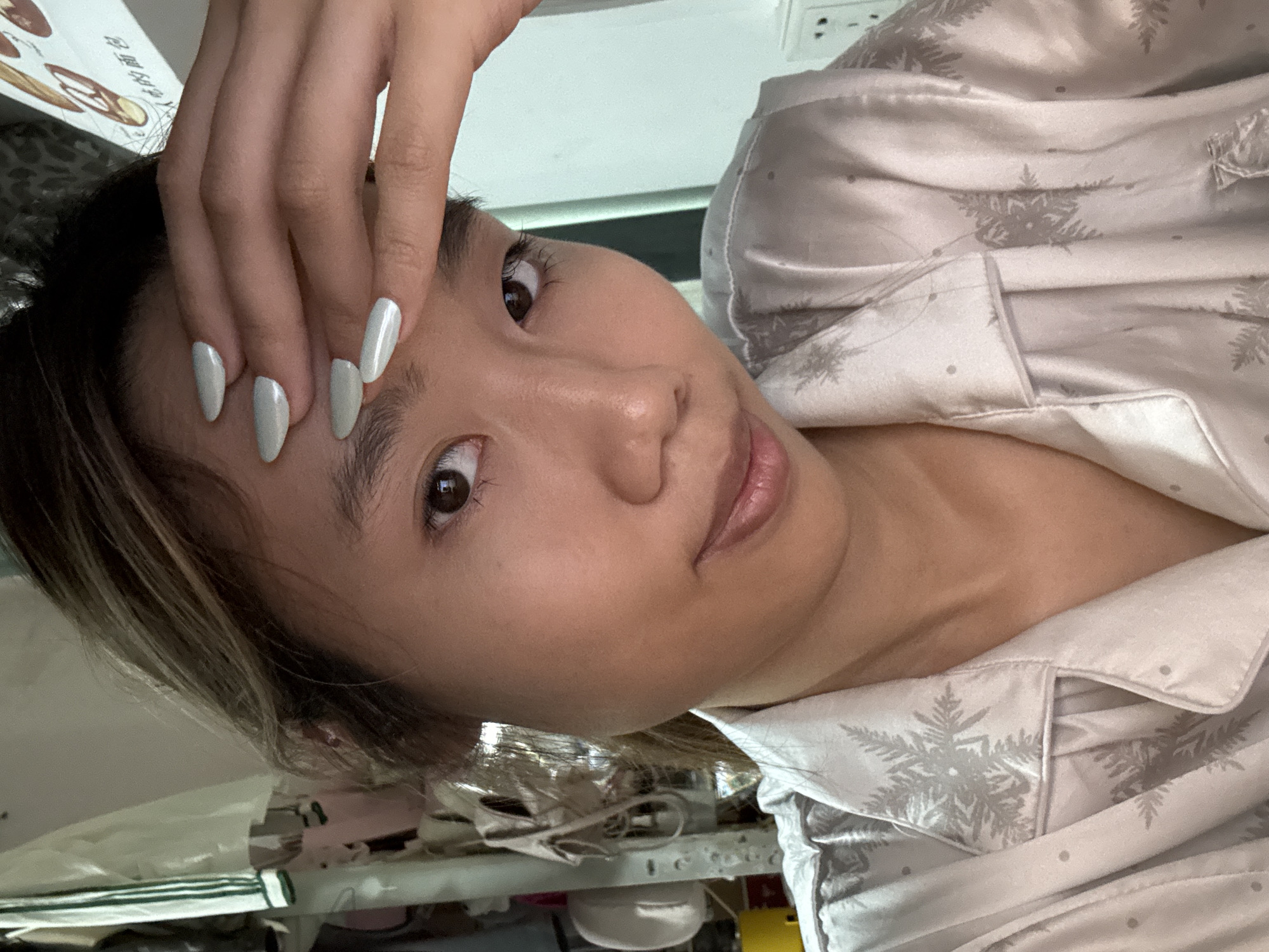 A selfie of a woman wearing light-colored pajamas with a snowflake pattern. She is holding her hand to her forehead, displaying pale-colored nails. The environment appears to be an indoor setting. The image does not provide information about the location being in Nan'an District, China.